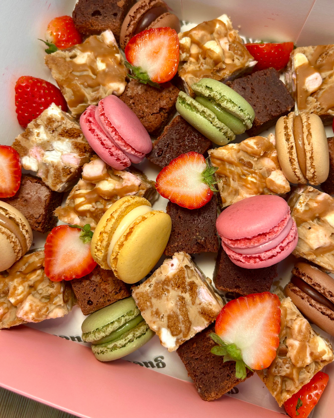 Assorted desserts including macarons, brownie squares, strawberry halves, and creamy caramel-topped bars