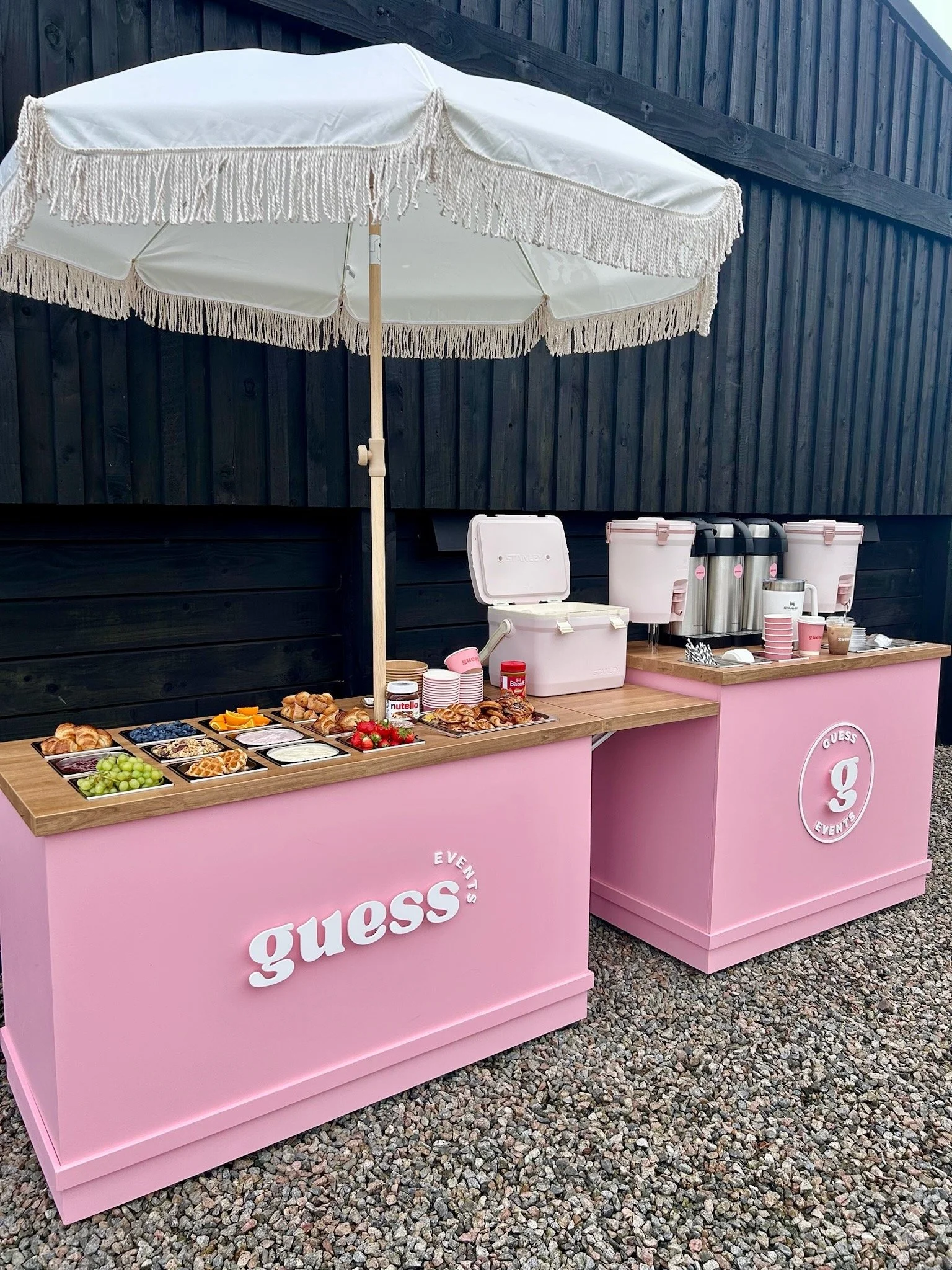 A pink outdoor refreshment stand with the GUESS Events logo, offering breakfast items such as croissants, blueberries, grapes, strawberries, and various beverages under a large white umbrella with fringe, set against a black wooden fence and gravel ground.