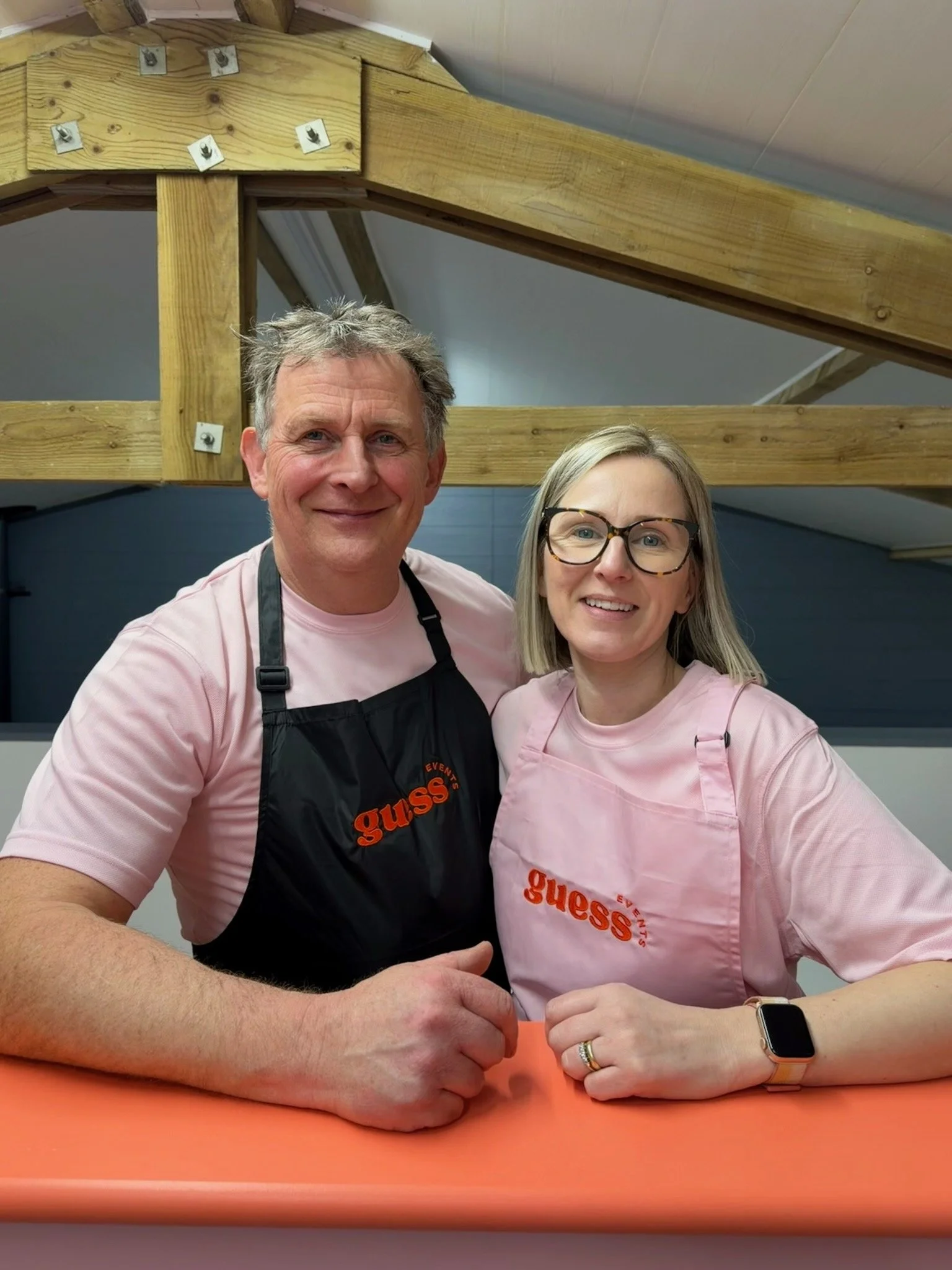 A smiling man and woman in pink shirts and black aprons with 'guess' logos, standing together in a room with wooden beams, posing for a photo.