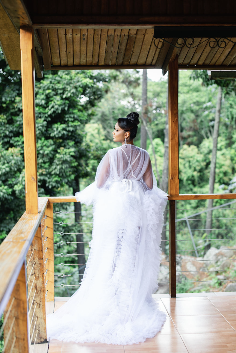 Woman in a wedding dress standing on a wooden balcony overlooking a lush green forest.