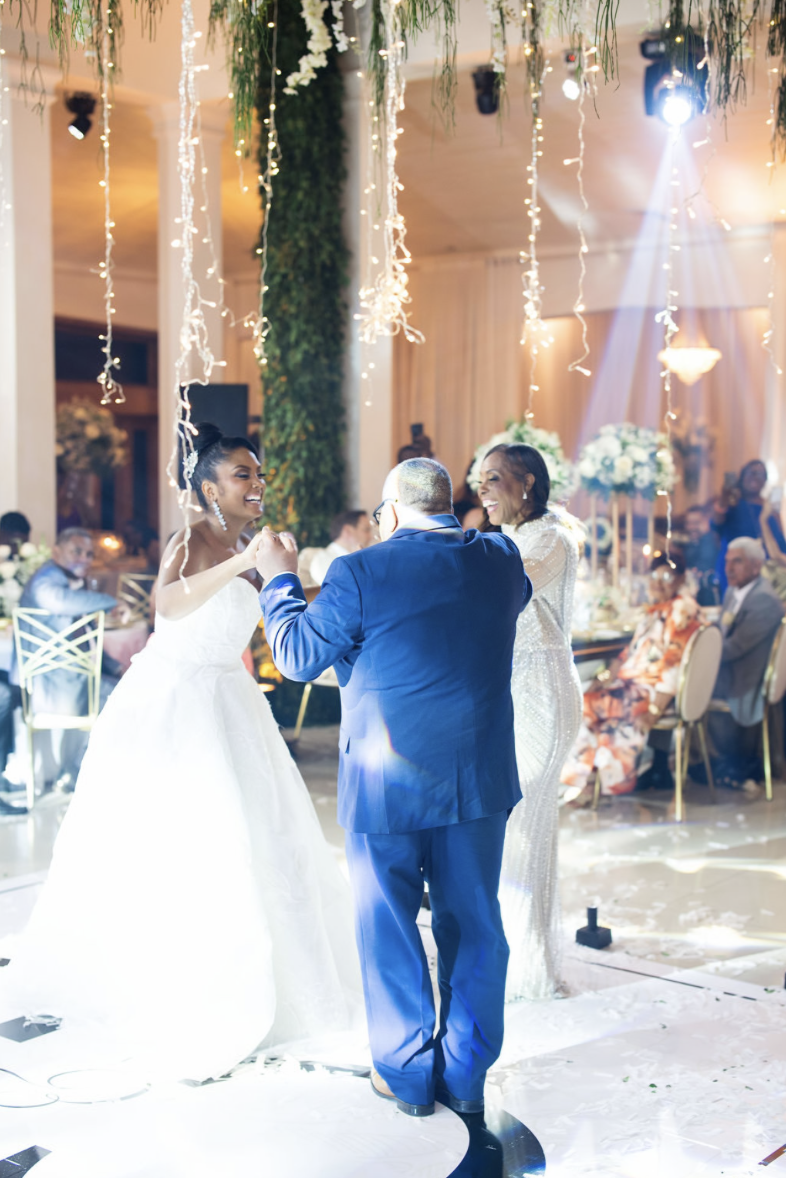 Bride and groom dancing at a wedding reception, surrounded by guests, with hanging fairy lights and floral decorations.