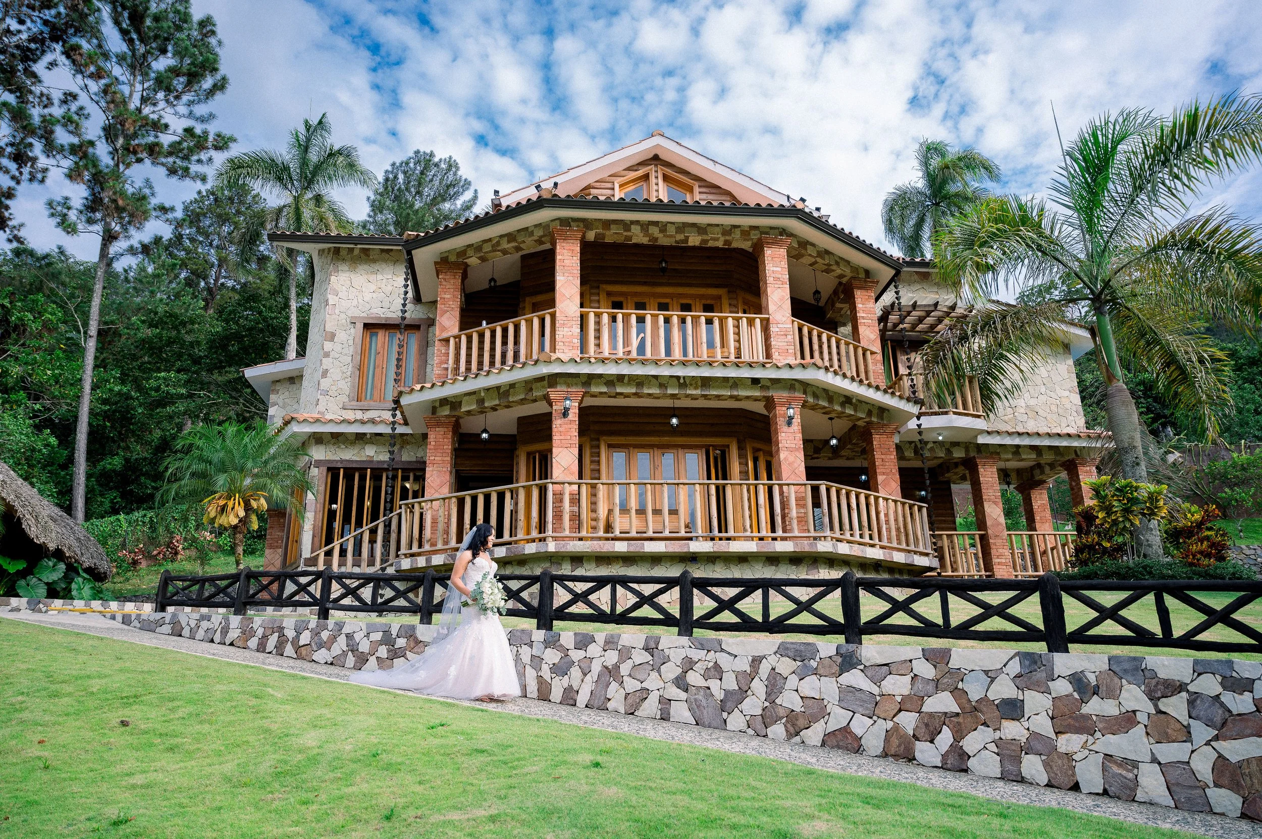 A bride in a wedding dress holding a bouquet walks on a grassy lawn in front of a large, multi-story house with wooden balconies, brick and stone exterior, surrounded by palm trees and lush vegetation under a partly cloudy sky.