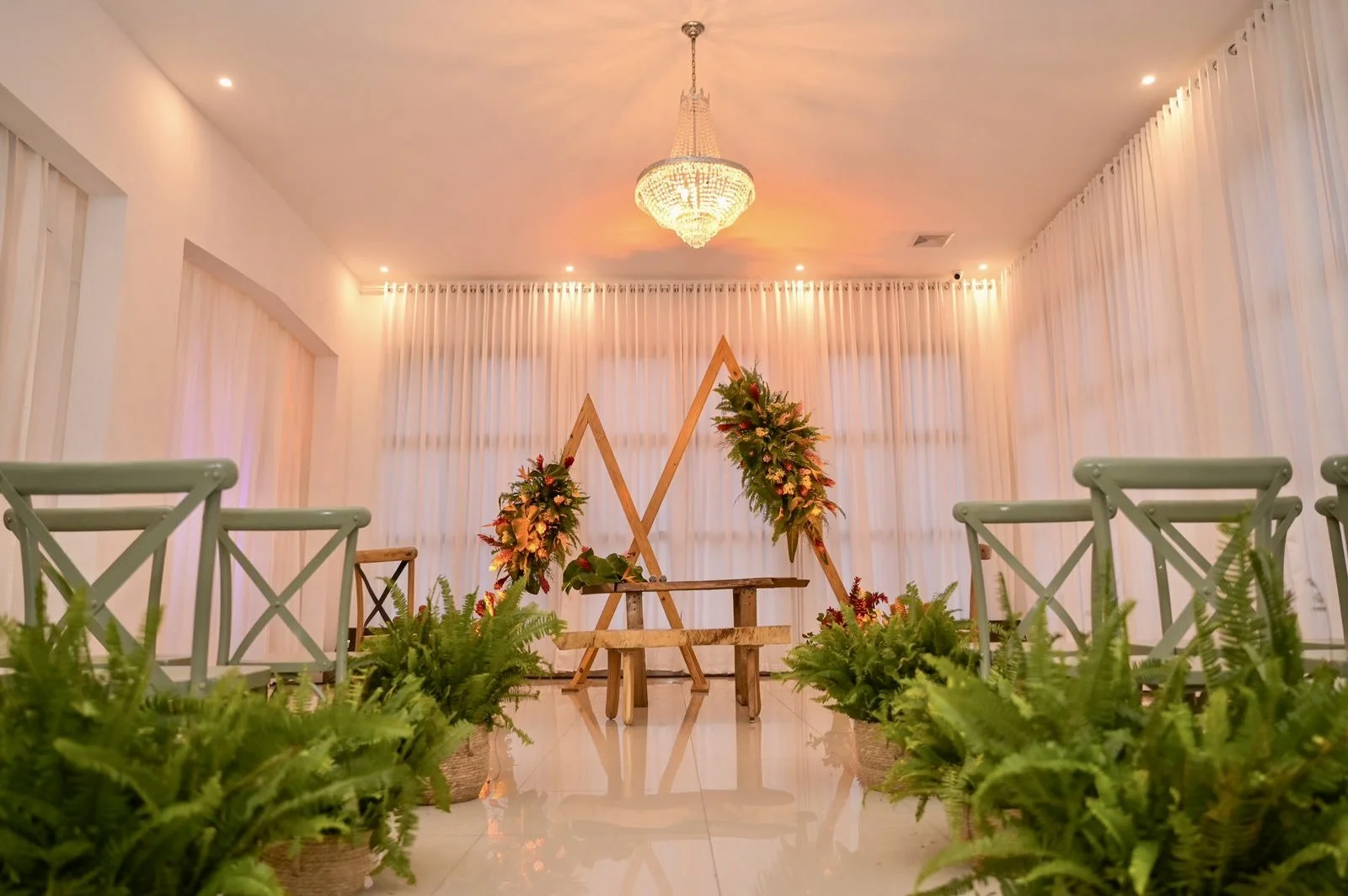Wedding ceremony setup with wooden chairs, floral arrangements, and a backdrop with an X-shaped decor under a chandelier in a well-lit room with white curtains.