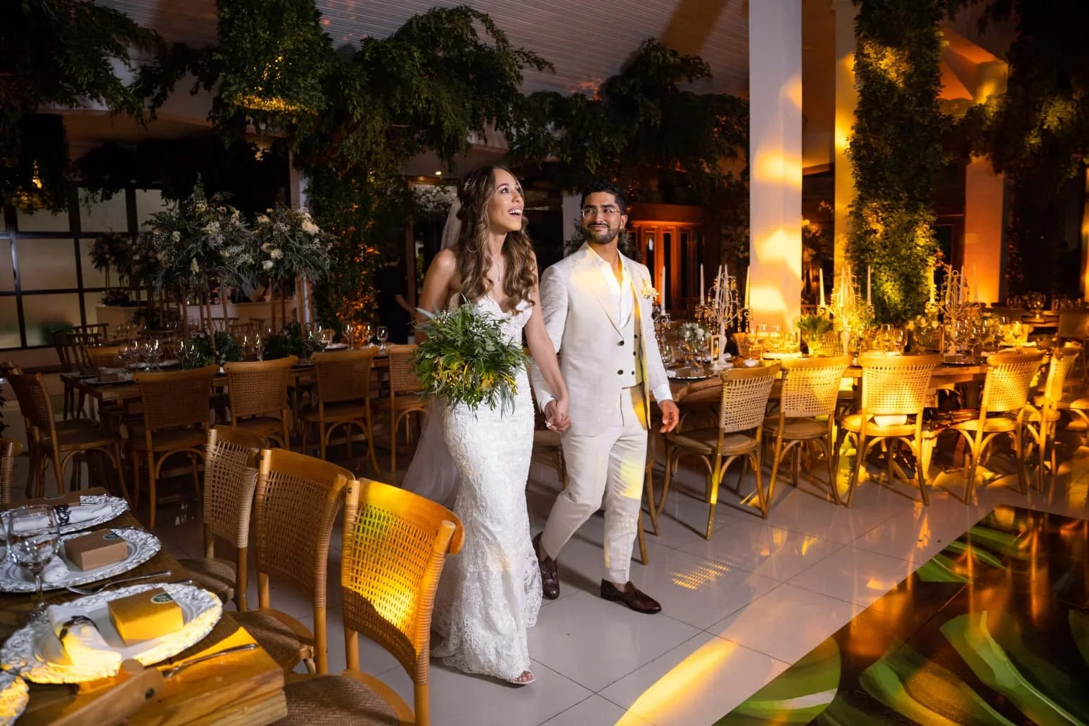 A bride and groom holding hands and smiling at their wedding reception, surrounded by decorated tables with floral arrangements and warm lighting.