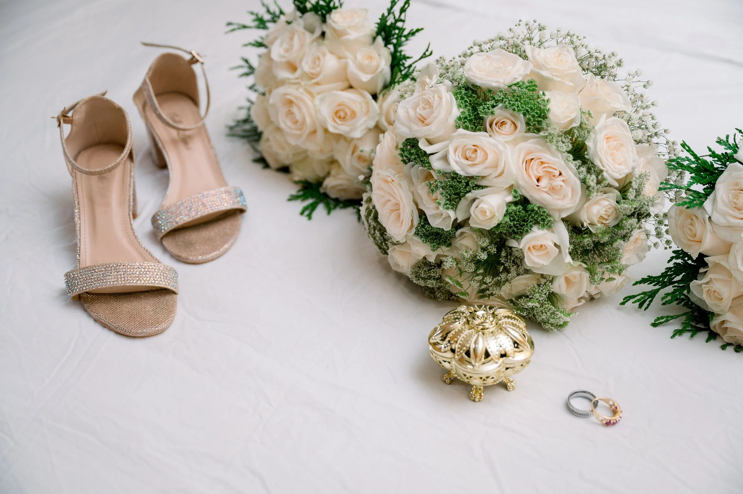 A pair of beige high-heeled sandals with rhinestone straps, two bouquets of white roses, two wedding rings, and a decorative gold box on a white surface.