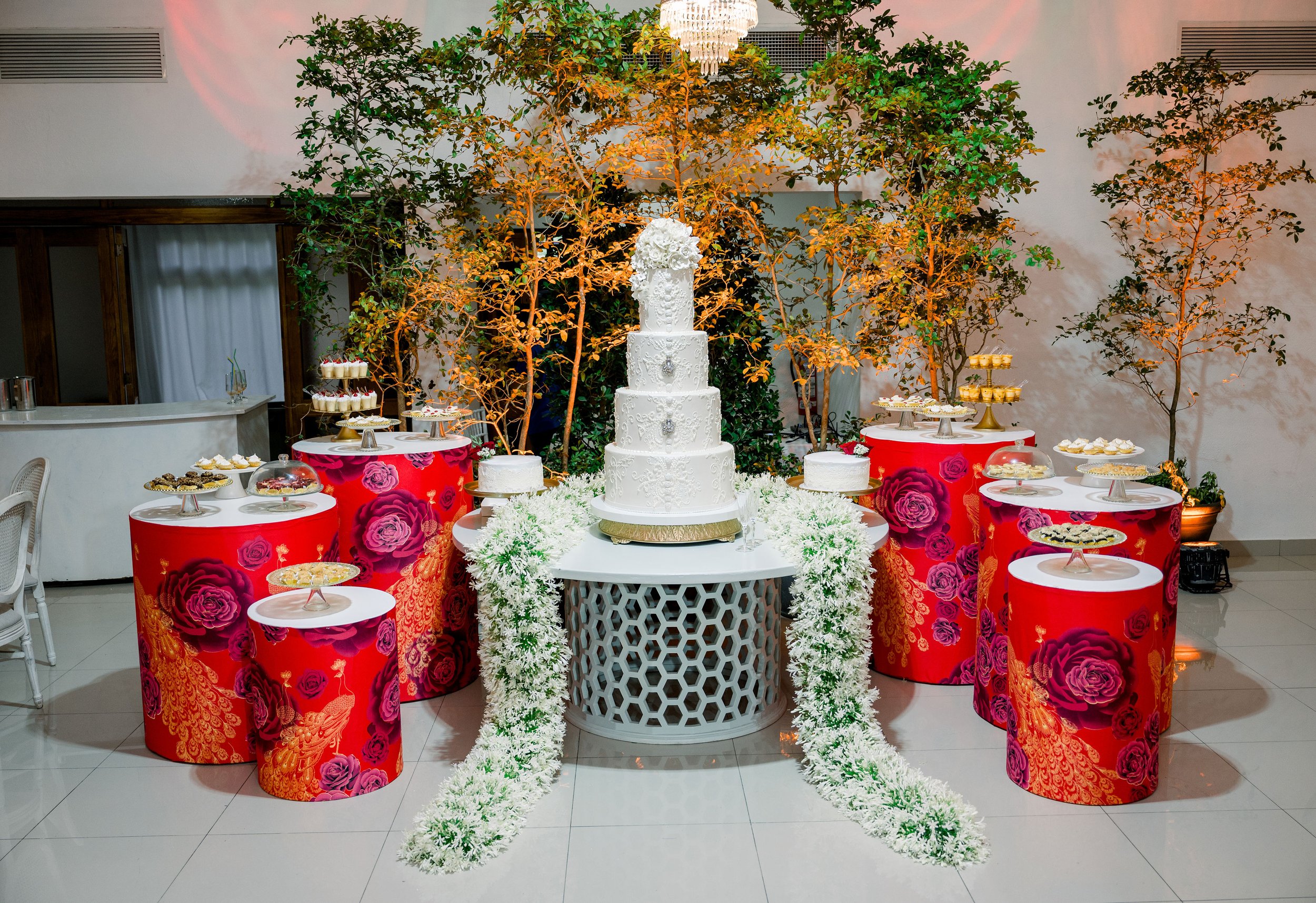 An elegant wedding cake display featuring a tall, multi-tiered white cake with intricate decorative patterns, surrounded by floral arrangements. The backdrop includes several small tables with desserts and treats, with red fabric covers decorated wit