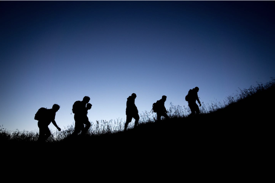 Silhouettes of five hikers walking uphill during dusk or dawn with backpacks.