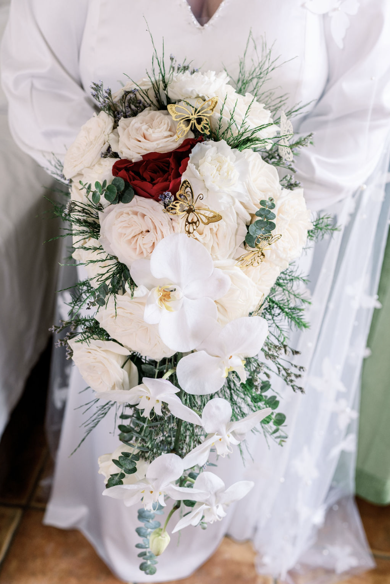A person holding a cascading wedding bouquet of white, blush, and deep red roses, white orchids, greenery, and decorative gold butterfly accents.