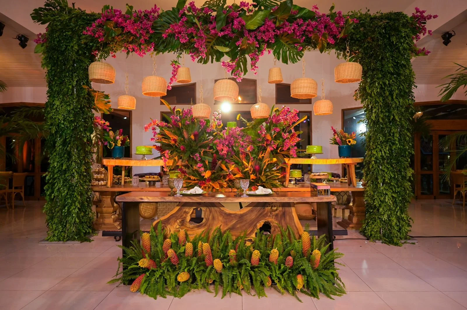Decorative indoor event space with lush green foliage, vibrant pink flowers on top and on sides, wooden tables with floral arrangements, and hanging wicker pendant lights, creating a tropical-inspired setting.