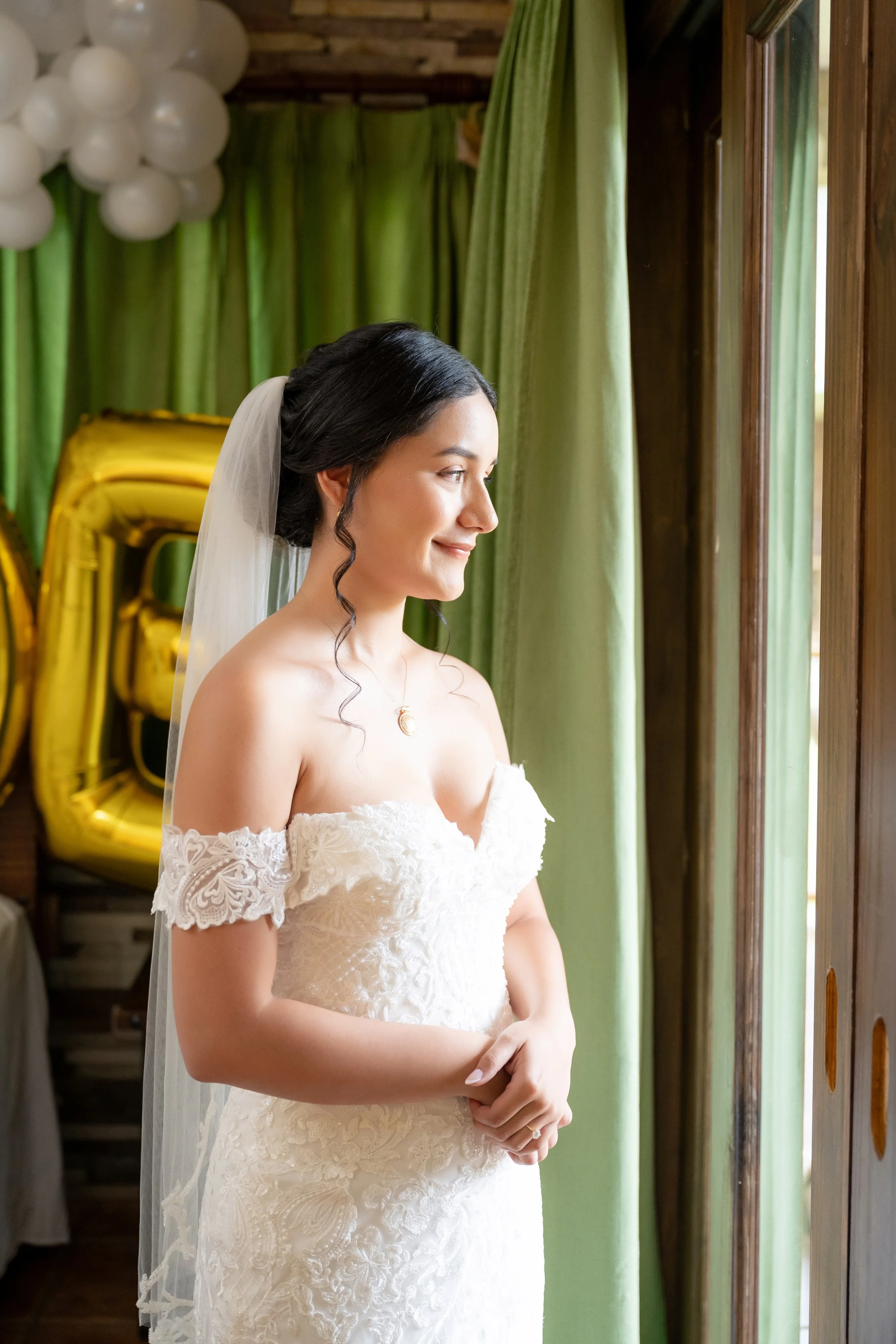 A bride with dark hair in an elegant white lace off-the-shoulder wedding dress stands near a window, smiling softly as she looks outside. She wears a veil and a delicate necklace, with balloons in the background.