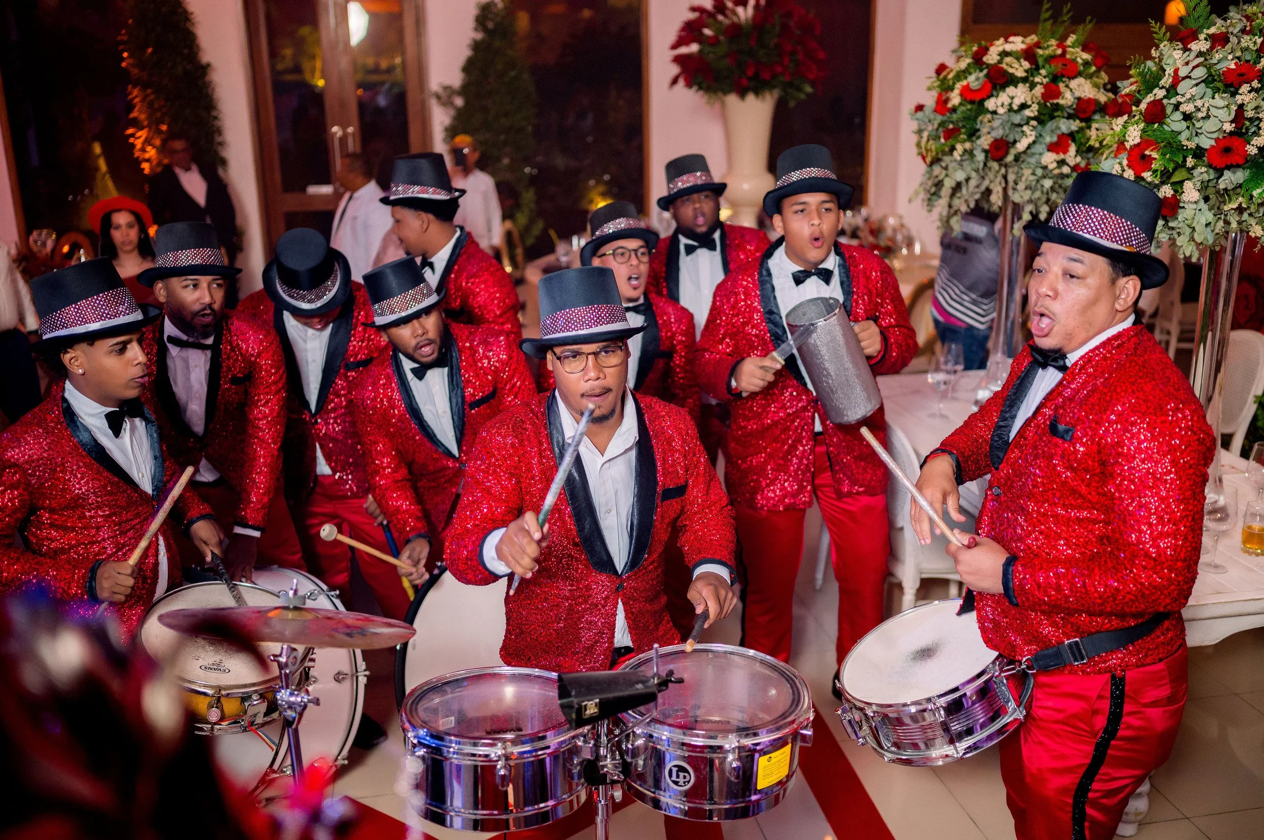 A group of musicians dressed in red sequined jackets, black top hats with red and white bands, white shirts, and black bow ties performing with drums and percussion instruments at an indoor event.