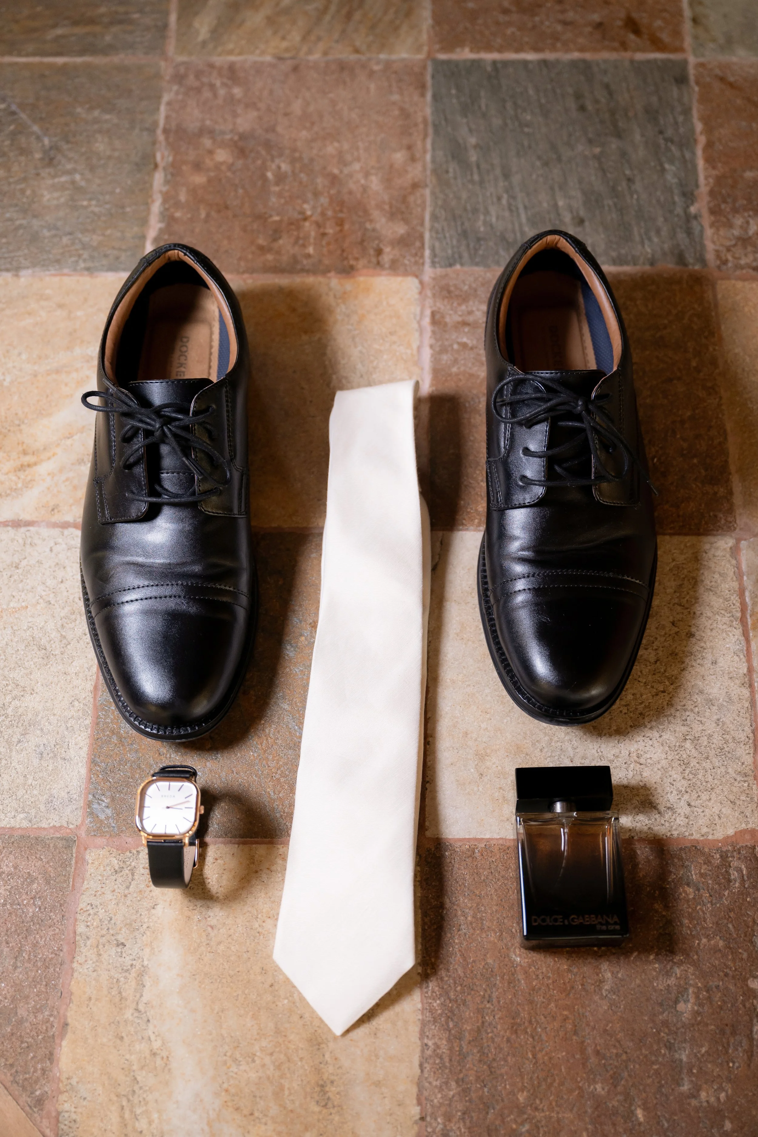 A pair of black leather dress shoes, a white tie, a wristwatch with a black strap, and a bottle of Dolce & Gabbana perfume on a tiled floor.