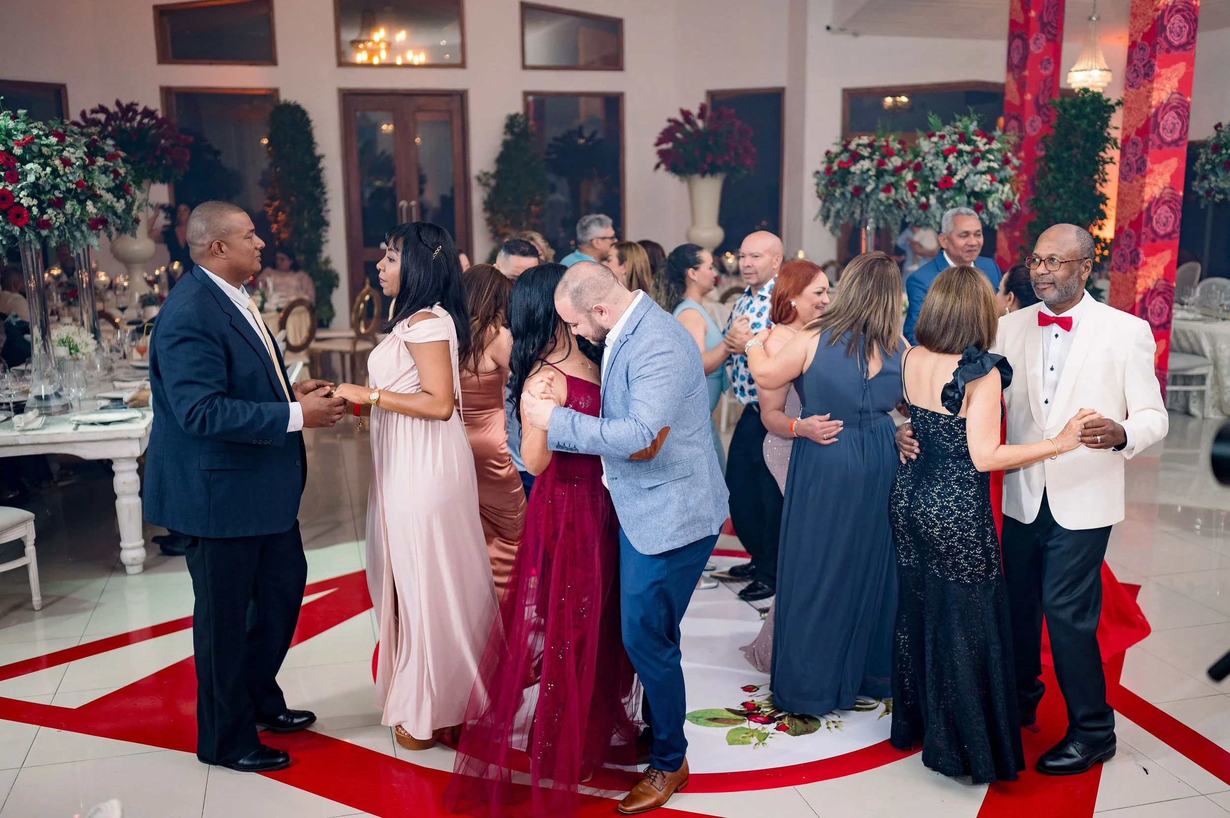People dancing and socializing at a wedding reception or celebration in a decorated indoor venue with floral arrangements and elegant decor.