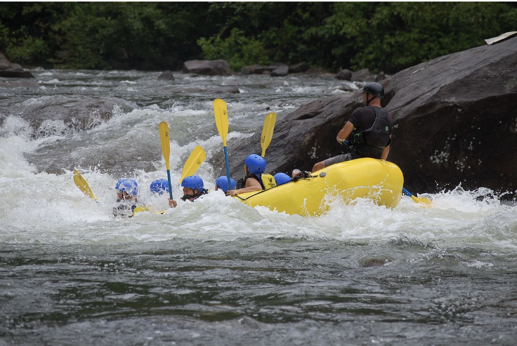 A group of people in a yellow raft navigating through whitewater rapids on a river, with an instructor steering them from the back.