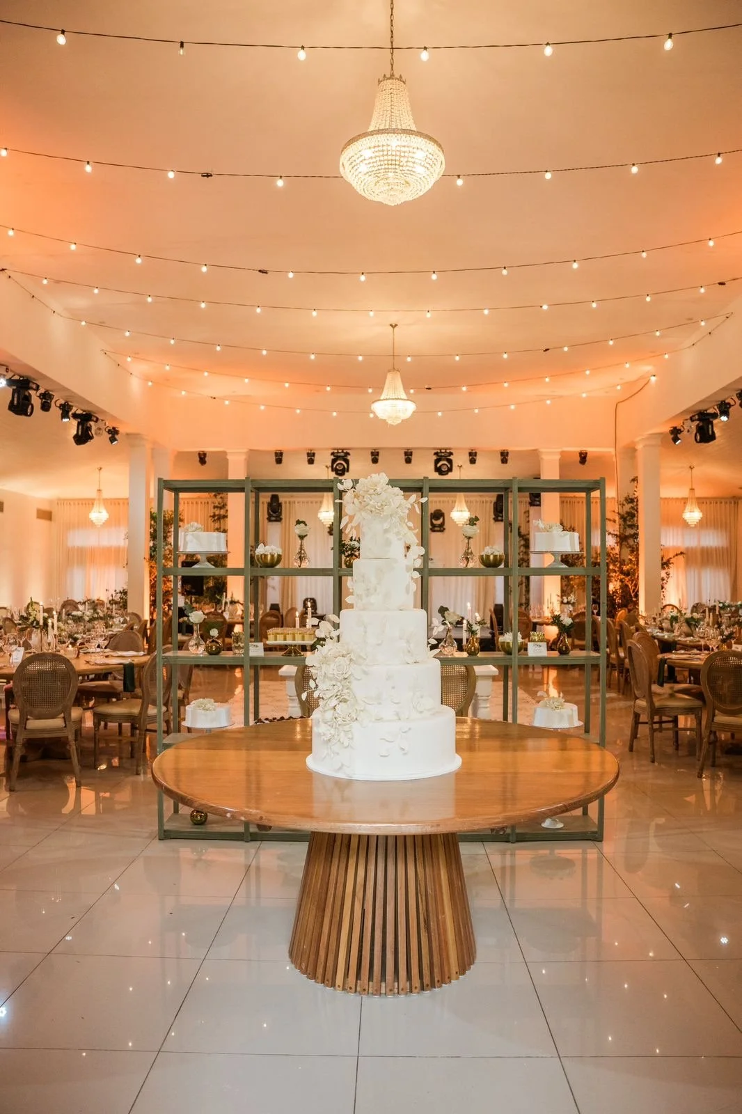 A tall, tiered white wedding cake decorated with flowers on a round wooden table in a decorated banquet hall with string lights and chandeliers.