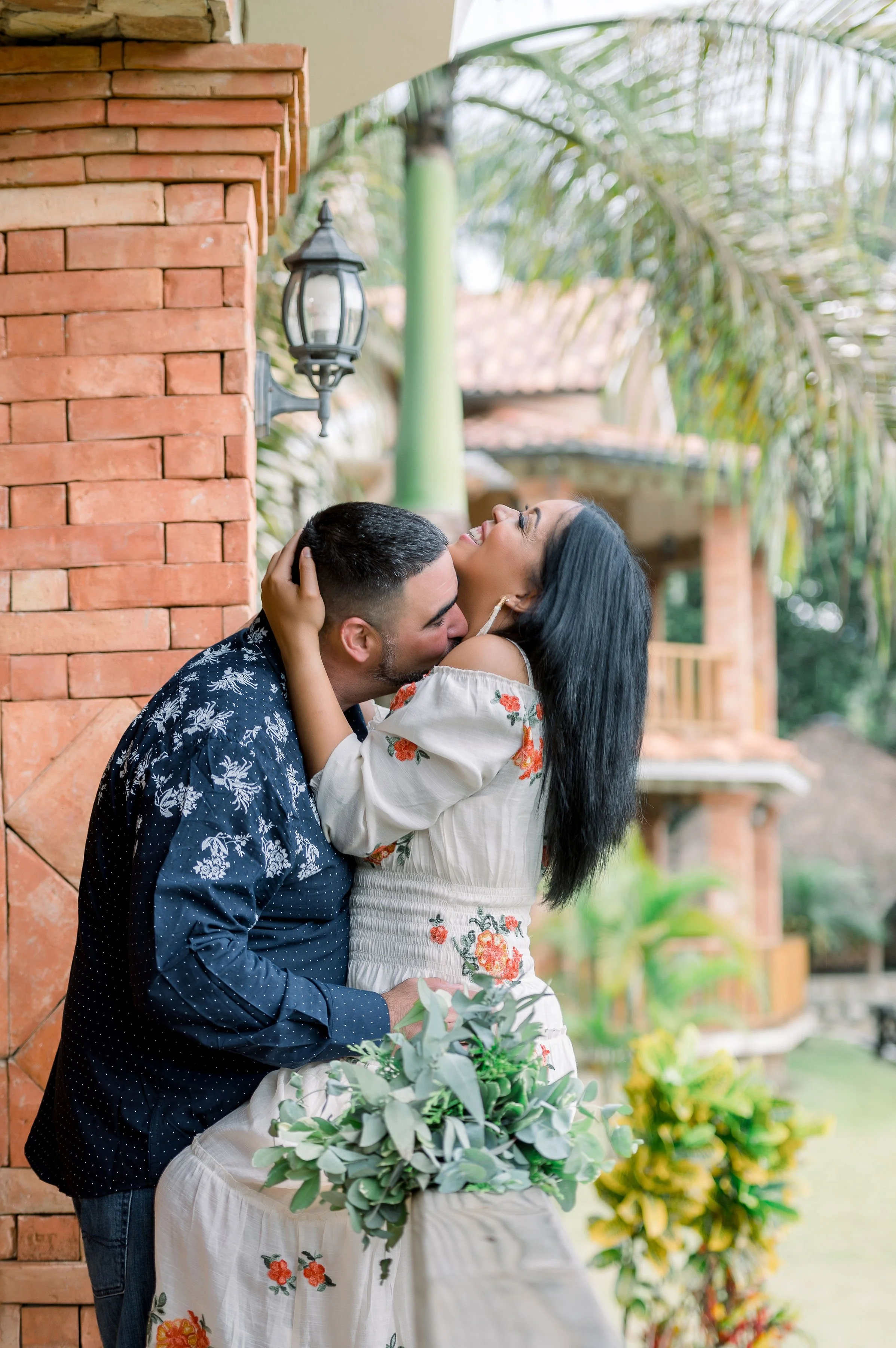 A couple sharing a joyful moment outdoors, with the man hugging the woman gently and she smiling and laughing. They are near a brick wall and surrounded by lush greenery and trees.