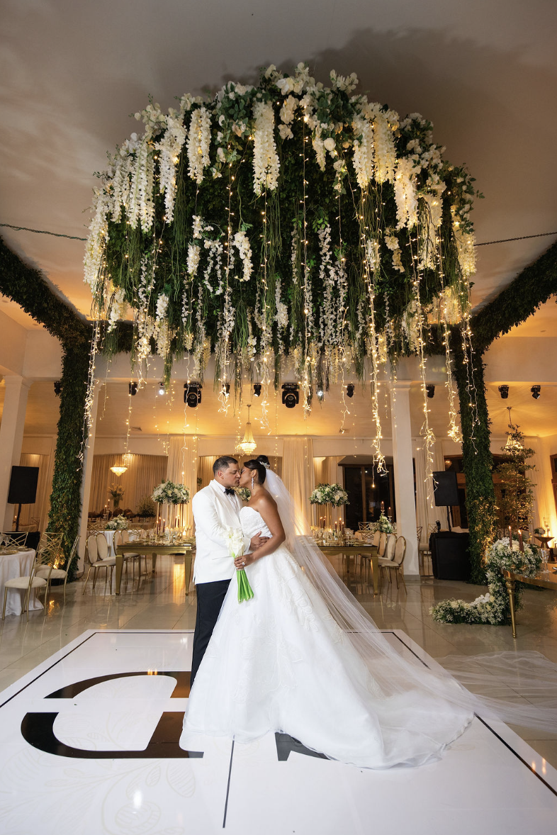 A bride and groom embrace on a dance floor at their wedding reception, with a large floral and light decorated canopy overhead in an elegant banquet hall.
