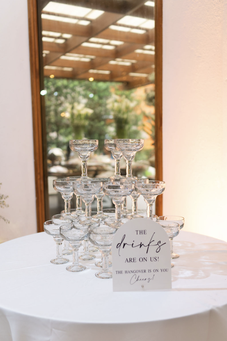 A champagne tower made of stacked coupe glasses on a round white table at a celebration, with a sign that reads 'The drinks are on us! The hangover is on you. Cheers!' behind the glasses, in front of a mirror reflecting an outdoor area.