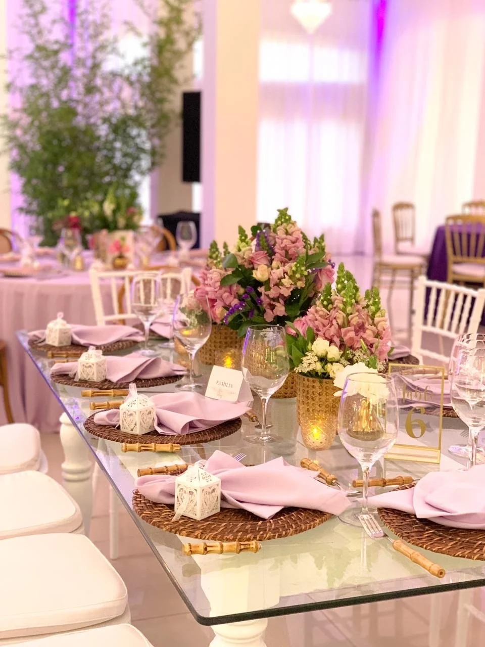 Elegant event table setup with pink floral centerpieces, pink napkins, white lantern decorations, and glassware on woven placemats.