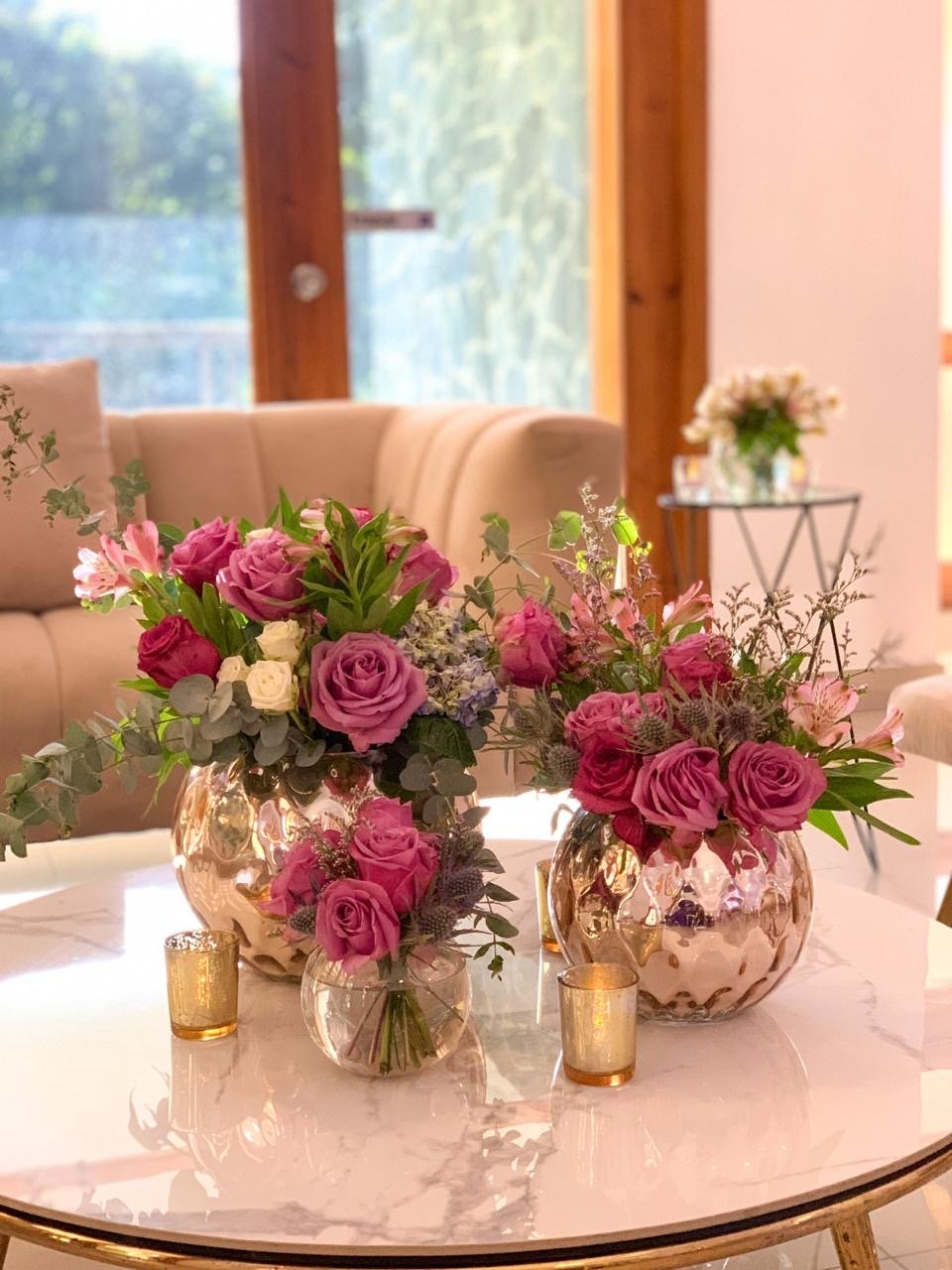 Two pink floral arrangements in gold vases on a white marble table, with small gold candle holders.
