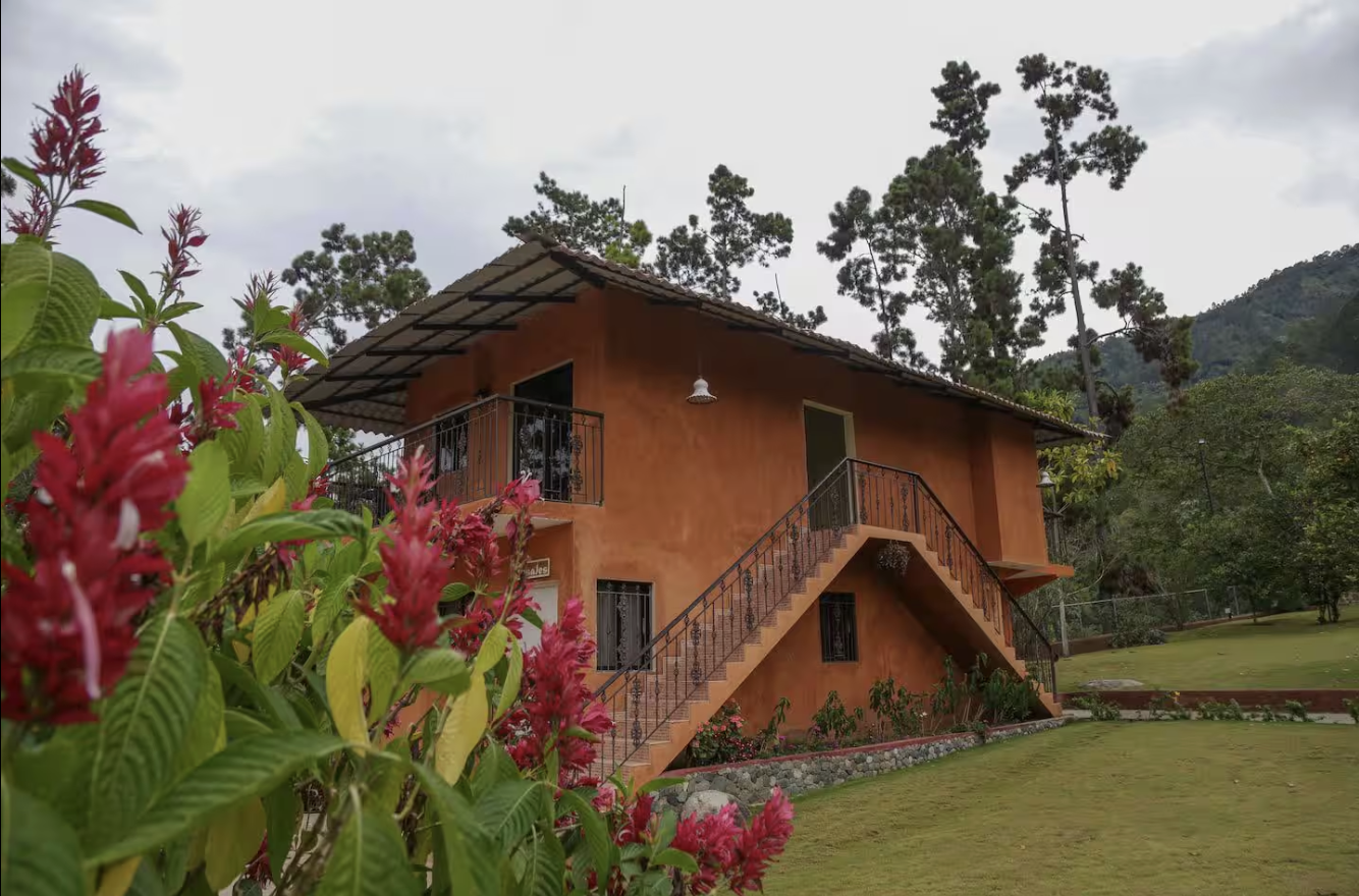 A two-story house with an orange stucco exterior, an outdoor staircase leading to the second floor, and surrounded by a lush green yard with trees and flowering plants.