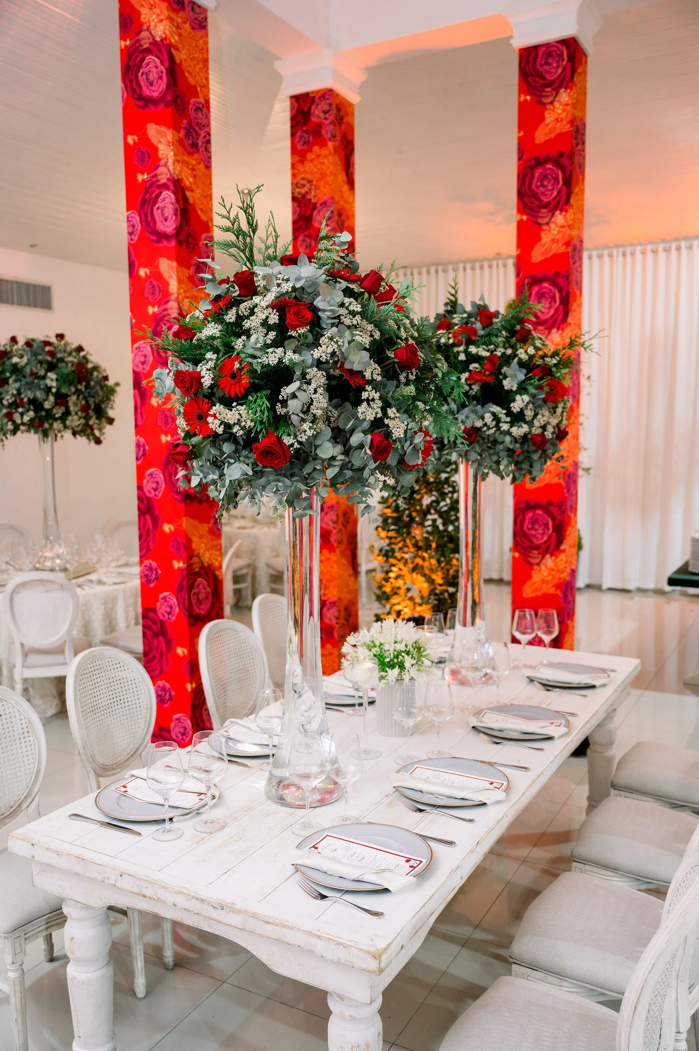 Elegant dining table set with white plates, silverware, and wine glasses, adorned with tall floral centerpieces of red roses, white baby's breath, and greenery, in a decorated event space with vibrant rose-patterned columns and white curtains.
