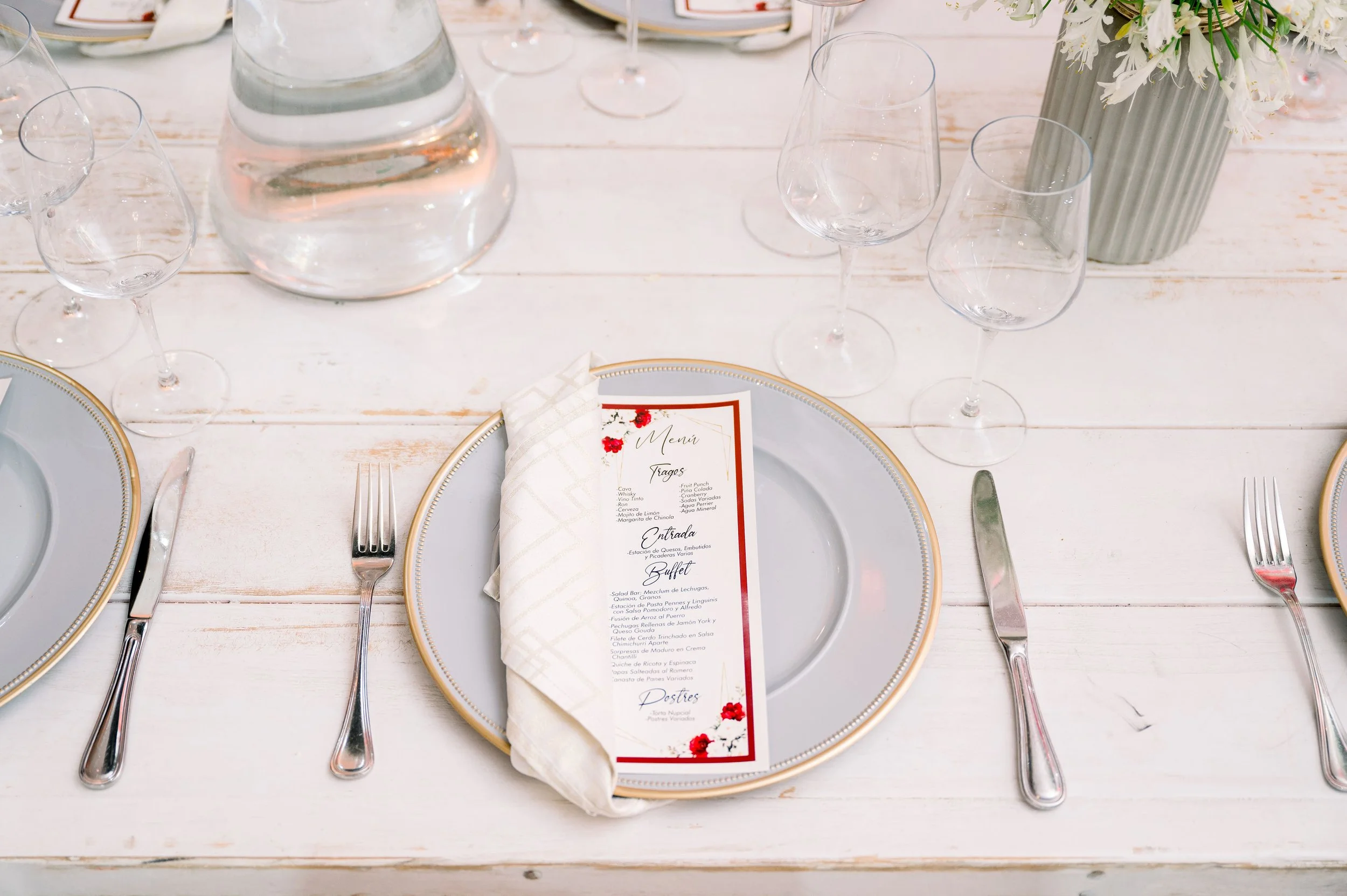Elegant place setting with a white plate with a gold rim, a folded cloth napkin, a menu card, and silverware arranged on a white wooden table. In the background, there are empty wine glasses, a vase with white flowers, and a water pitcher.