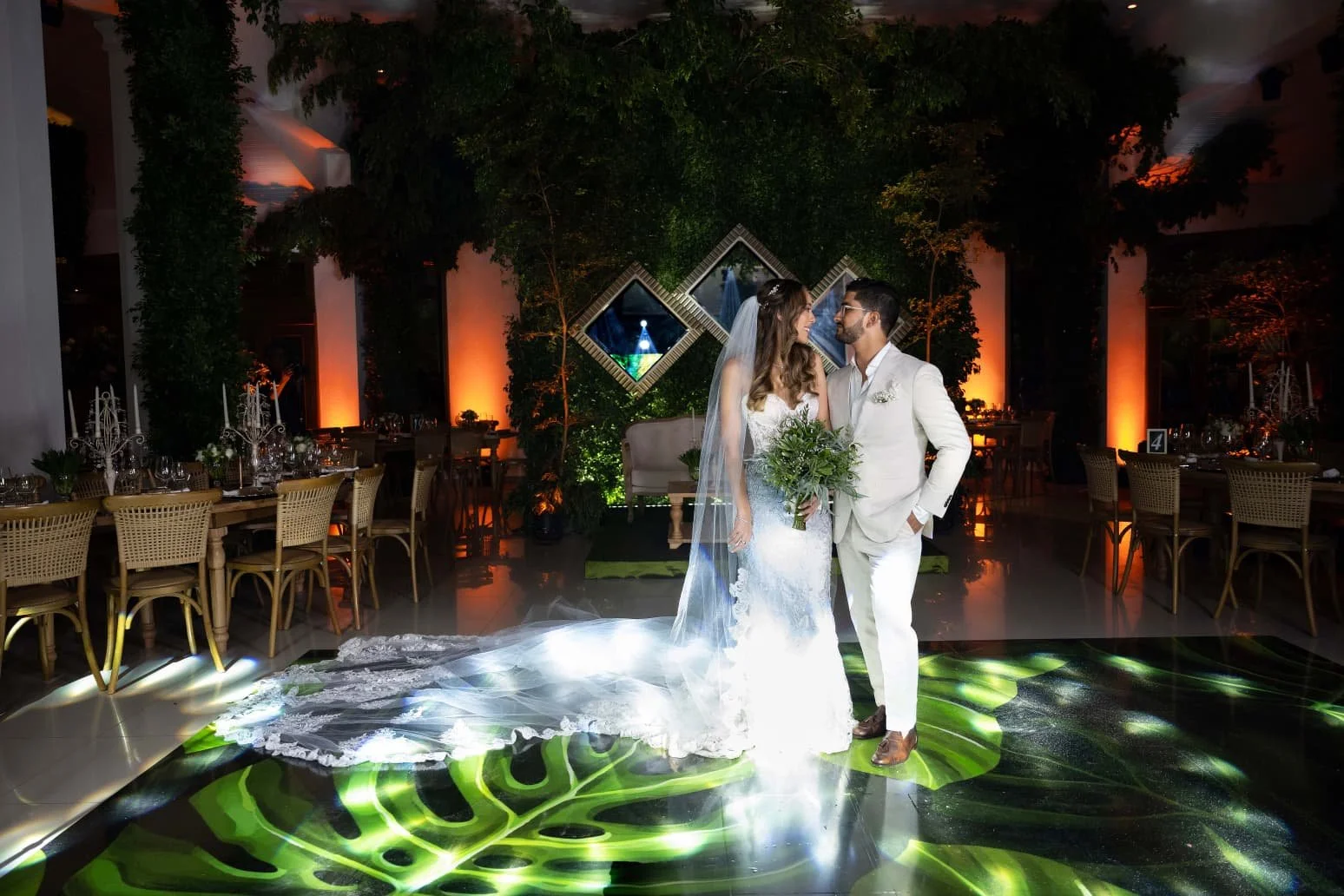 A bride and groom stand close together on a dance floor at their wedding reception, with the bride holding a bouquet of greenery, in a decorated indoor venue with warm lighting and framed wall art in the background.
