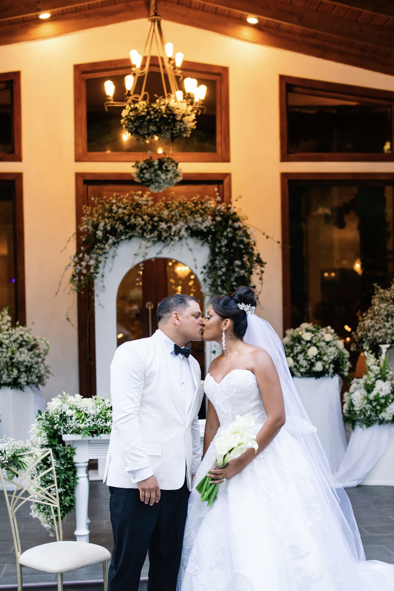 A bride and groom share a kiss at their wedding reception, with floral arrangements and a chandelier in the background.