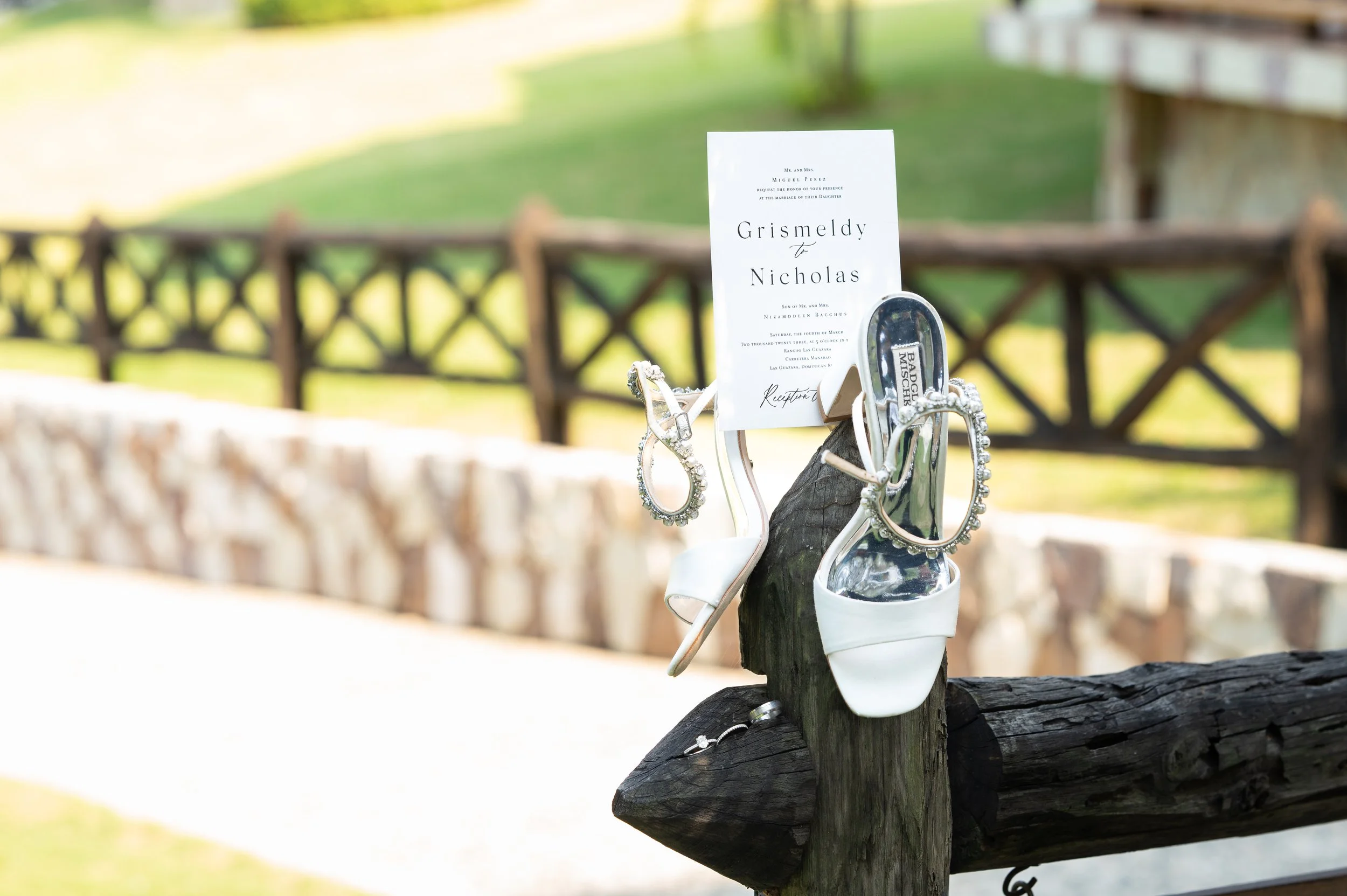 A wedding invitation is attached to a wooden post, topped with white high-heeled shoes and jewelry, with a blurred outdoor background of a fence and greenery.