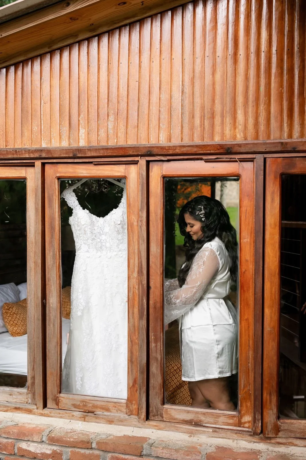 A woman in a white satin robe standing inside a wooden-framed glass door, looking at her wedding dress hanging inside.