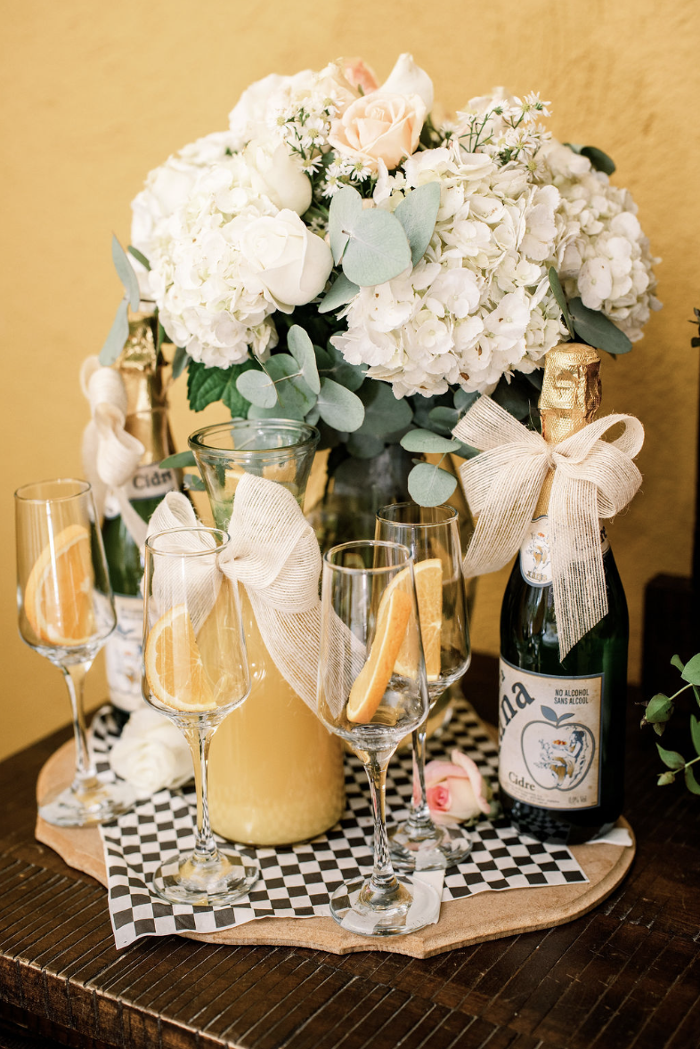 A floral arrangement with white and pale pink flowers, eucalyptus leaves, and a bow, alongside bottles and glasses of non-alcoholic cider with orange slices, on a wooden table.