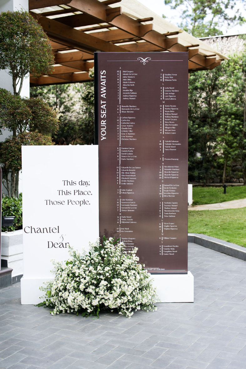 Wedding seating chart display with white floral arrangement in front of it, outdoors with trees and a wooden pergola.