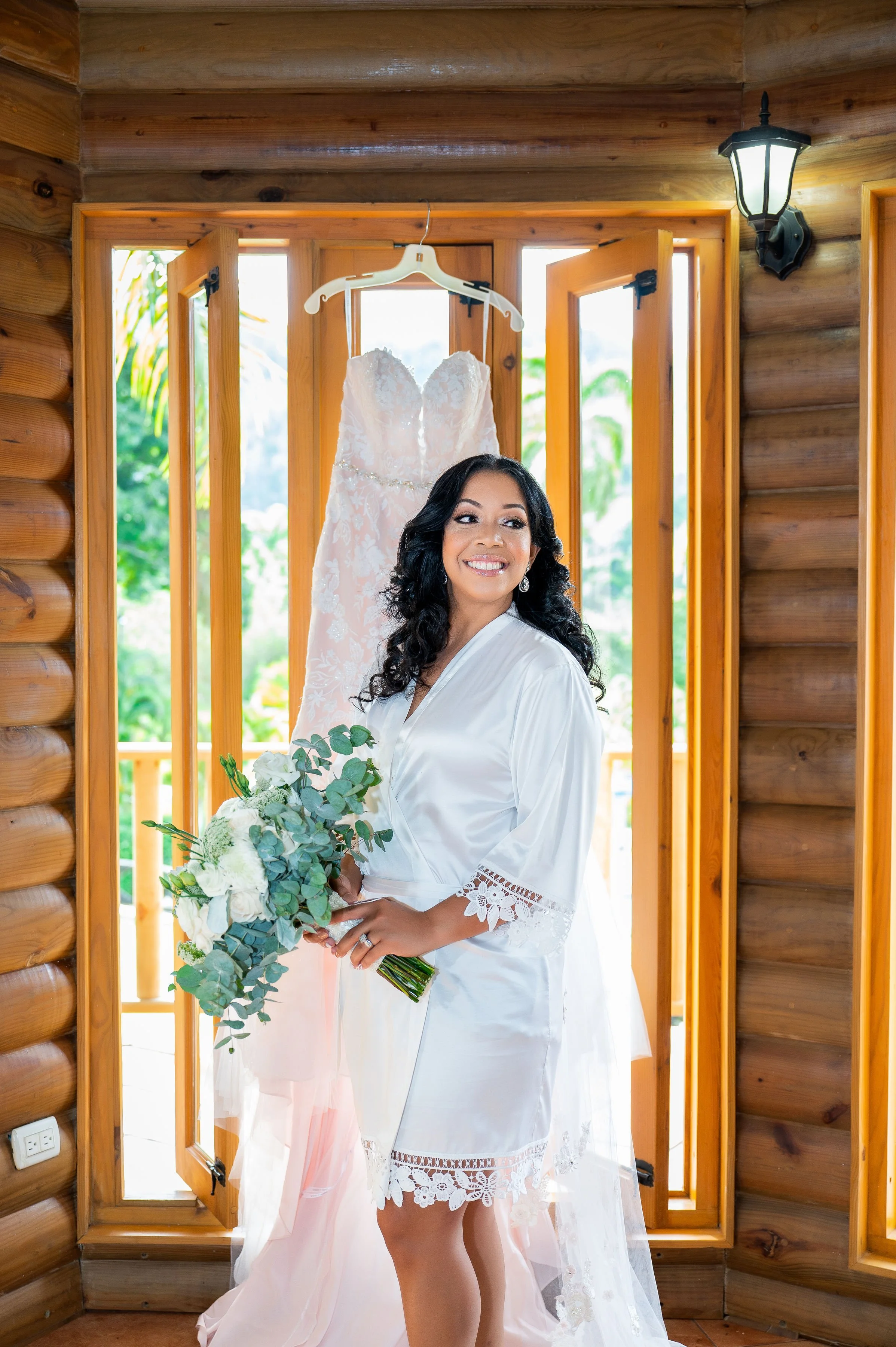 Smiling woman in white satin robe holding a bouquet of white and green flowers, standing in front of a wedding dress hanging in a wooden room with open windows.