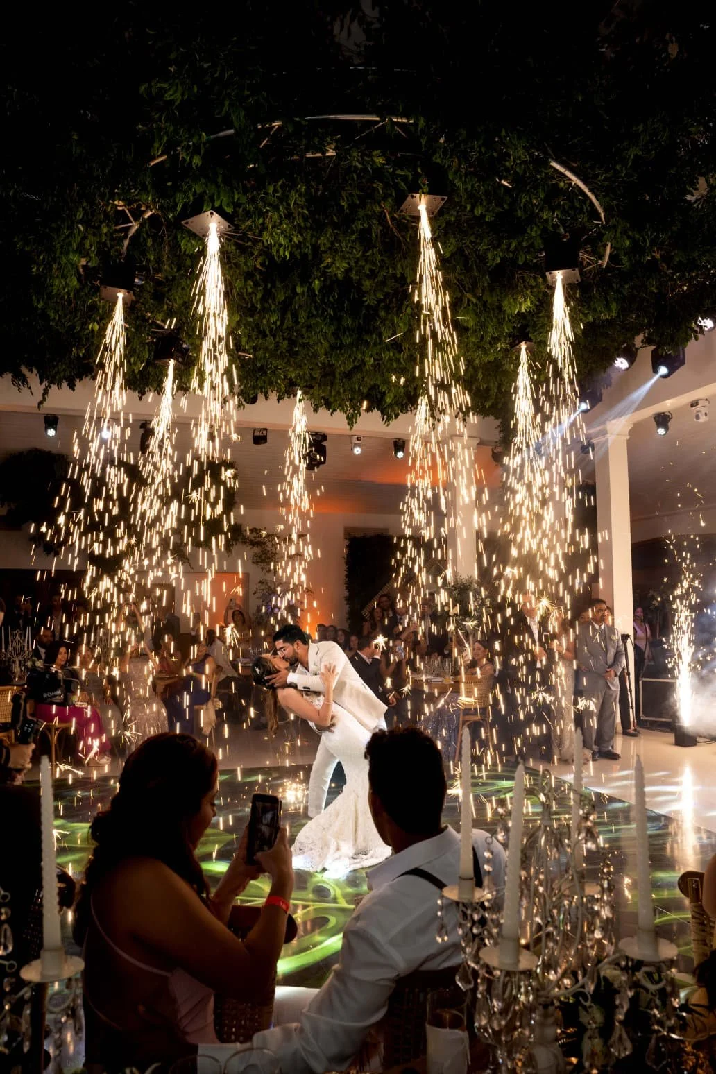 A bride and groom kissing during their wedding celebration inside a decorated venue with sparklers and fireworks, surrounded by guests and elegant decor.