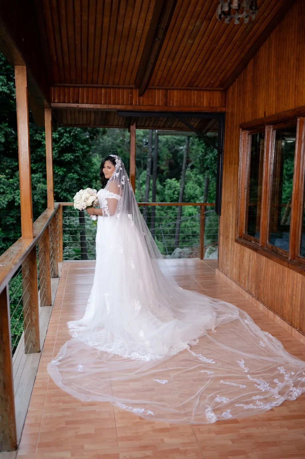 Bride in white wedding dress with lace details, holding a bouquet of white flowers, standing on a wooden balcony with a forest background.