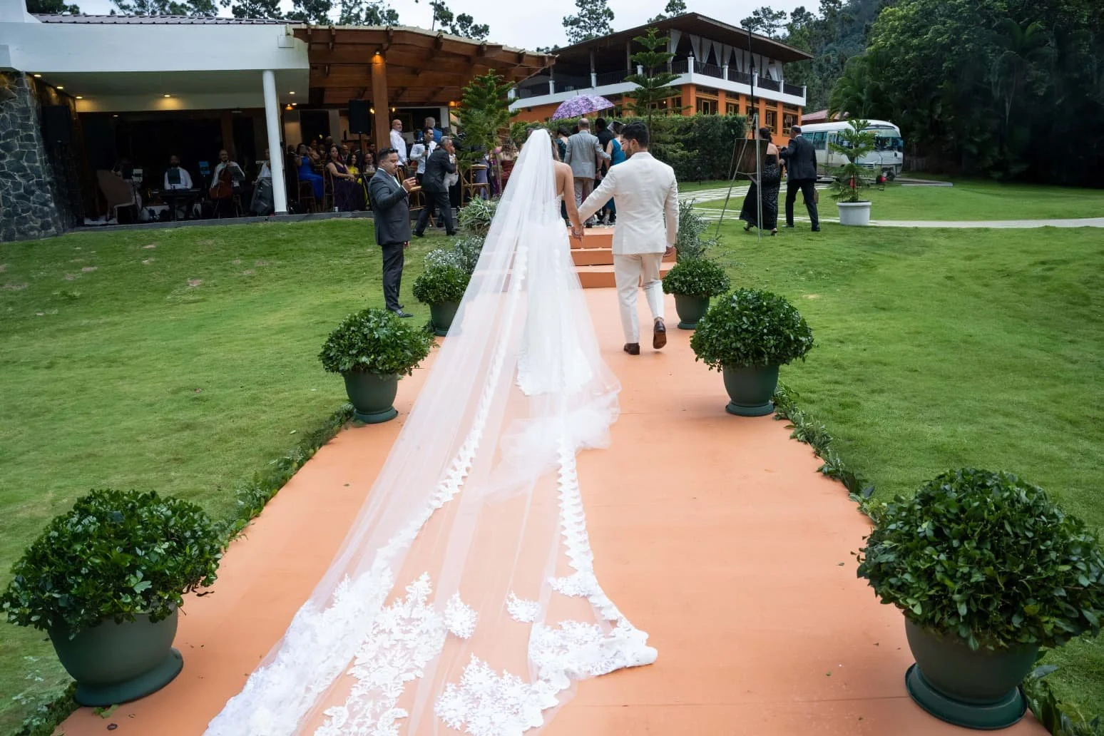 Bride and groom walking hand in hand down an outdoor aisle at a wedding, with guests seated in a venue in the background, and potted plants lining the aisle.
