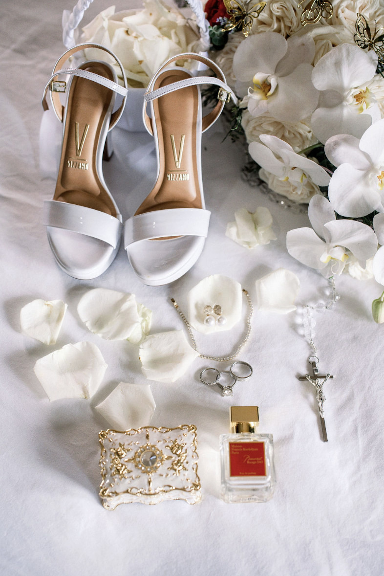 A pair of white high-heeled shoes with ankle straps, surrounded by white flowers, rose petals, jewelry including pearl earrings, a necklace, and rings, along with a perfume bottle on a white cloth.