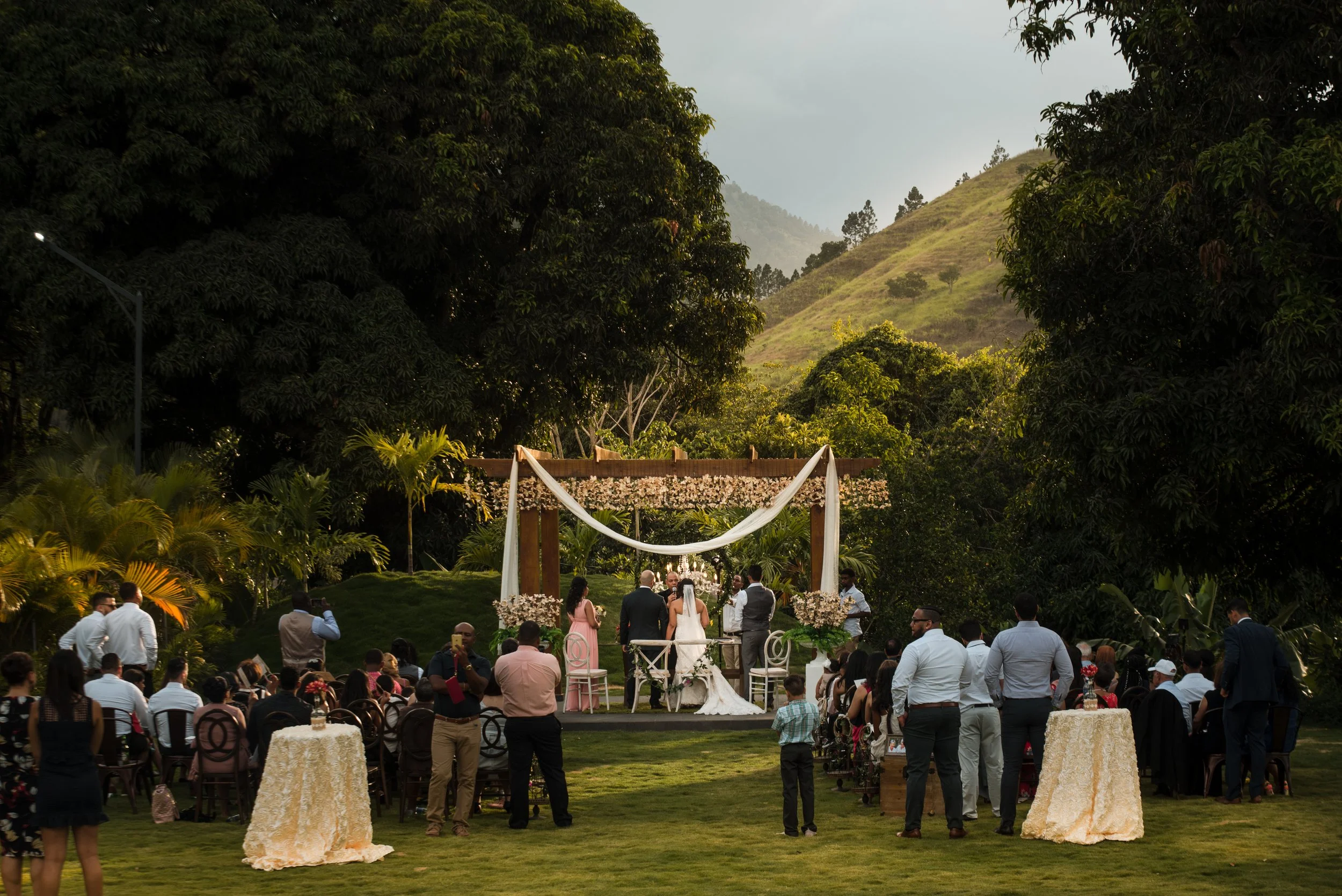 Outdoor wedding ceremony with guests seated on either side, officiant, bride, and groom standing under a decorated wooden arch in a lush garden setting with trees and mountains in the background.