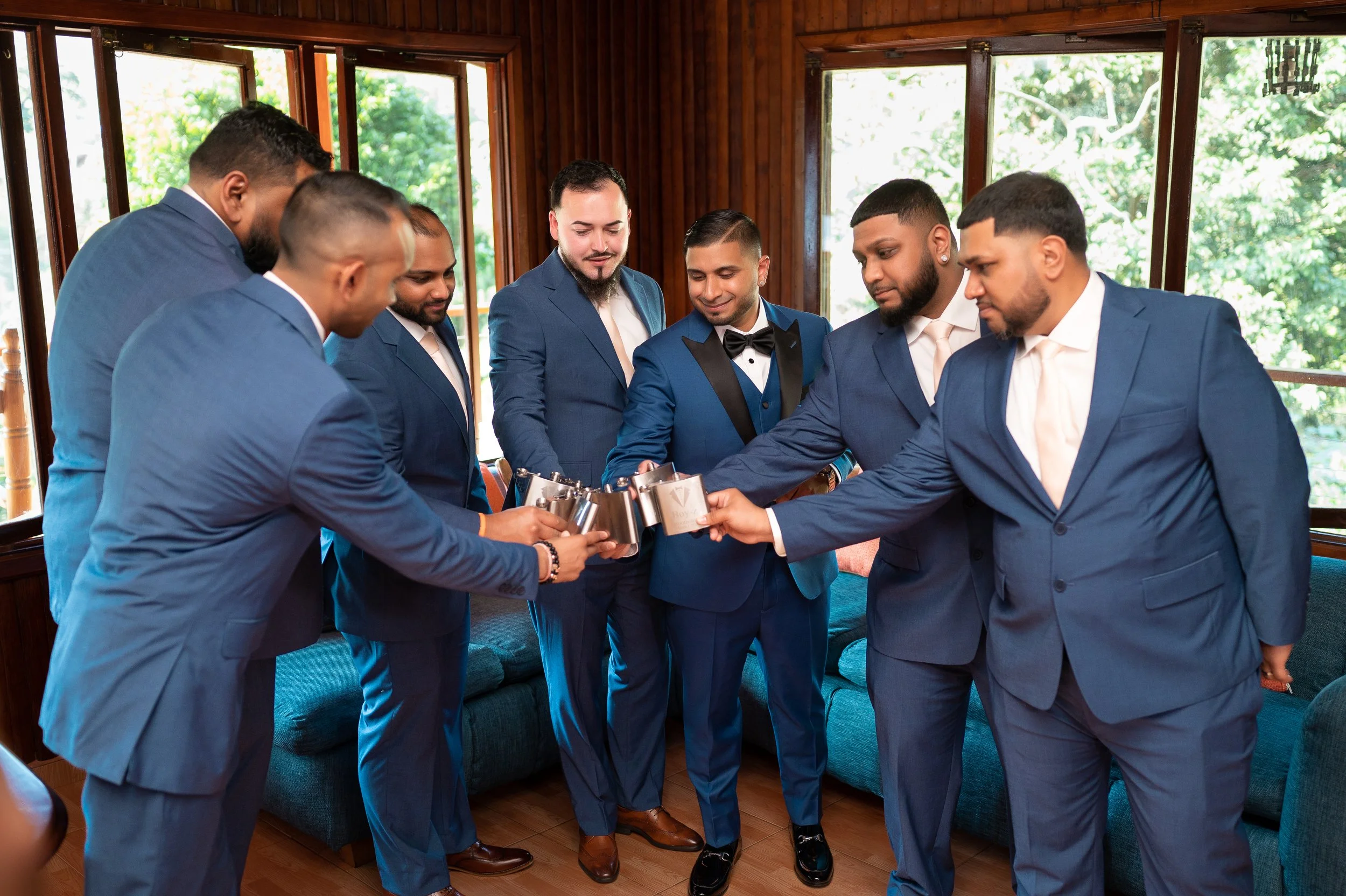 Group of seven men in blue suits and tuxedos clinking metallic mugs together in celebration indoors with large windows and greenery outside.