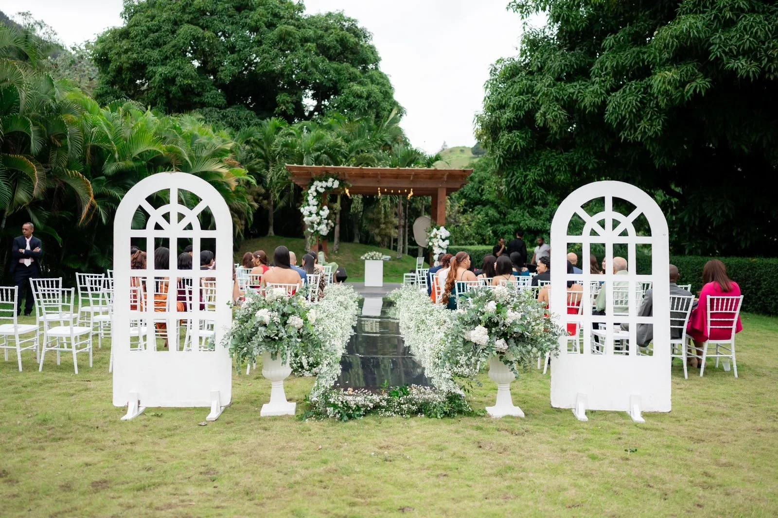 Outdoor wedding ceremony setup with white chairs, floral arrangements, and an archway on a grassy area surrounded by trees.