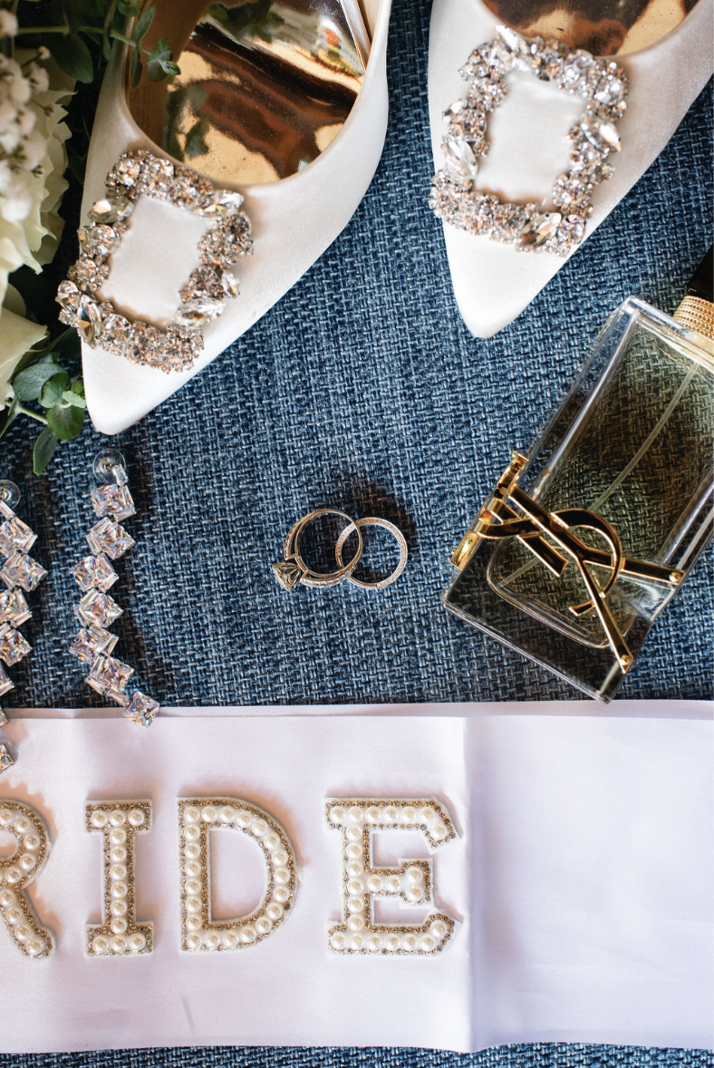 A collection of jewelry including rings, earrings, and a necklace, displayed on a textured blue fabric with a white cloth underneath. The jewelry has sparkling stones and pearls, and there is a metallic clutch purse and perfume bottle nearby. The wor