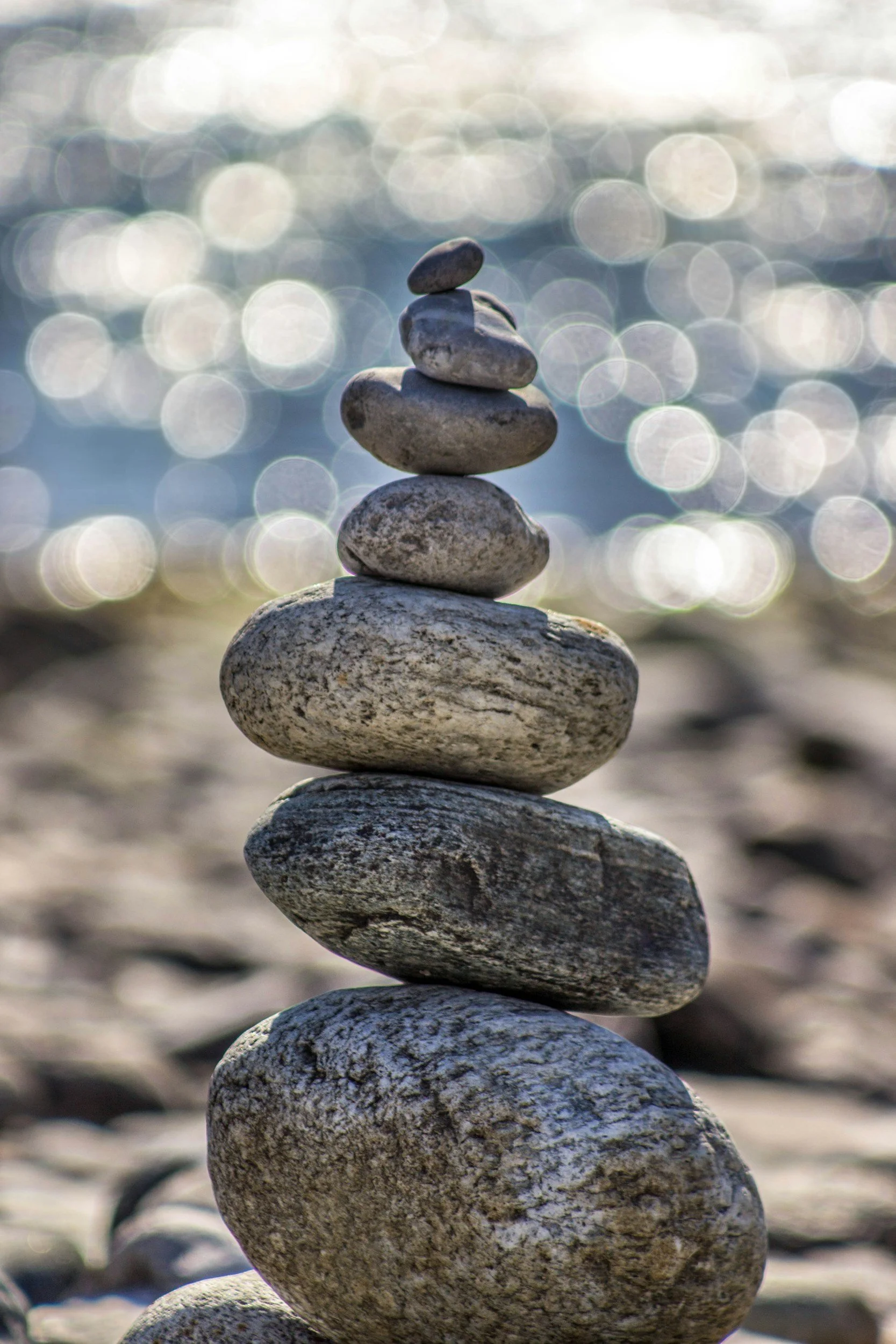 A stack of seven smooth rocks balanced on top of each other near a body of water with sunlight reflecting off the surface in the background.