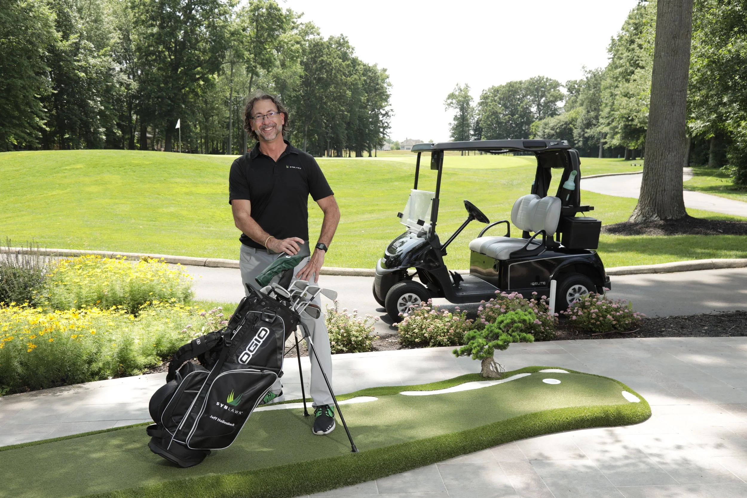 Putting green on patio with man standing on it behind a set of golf clubs, with a golf cart behind him and the 18th hold of a golf course in the background