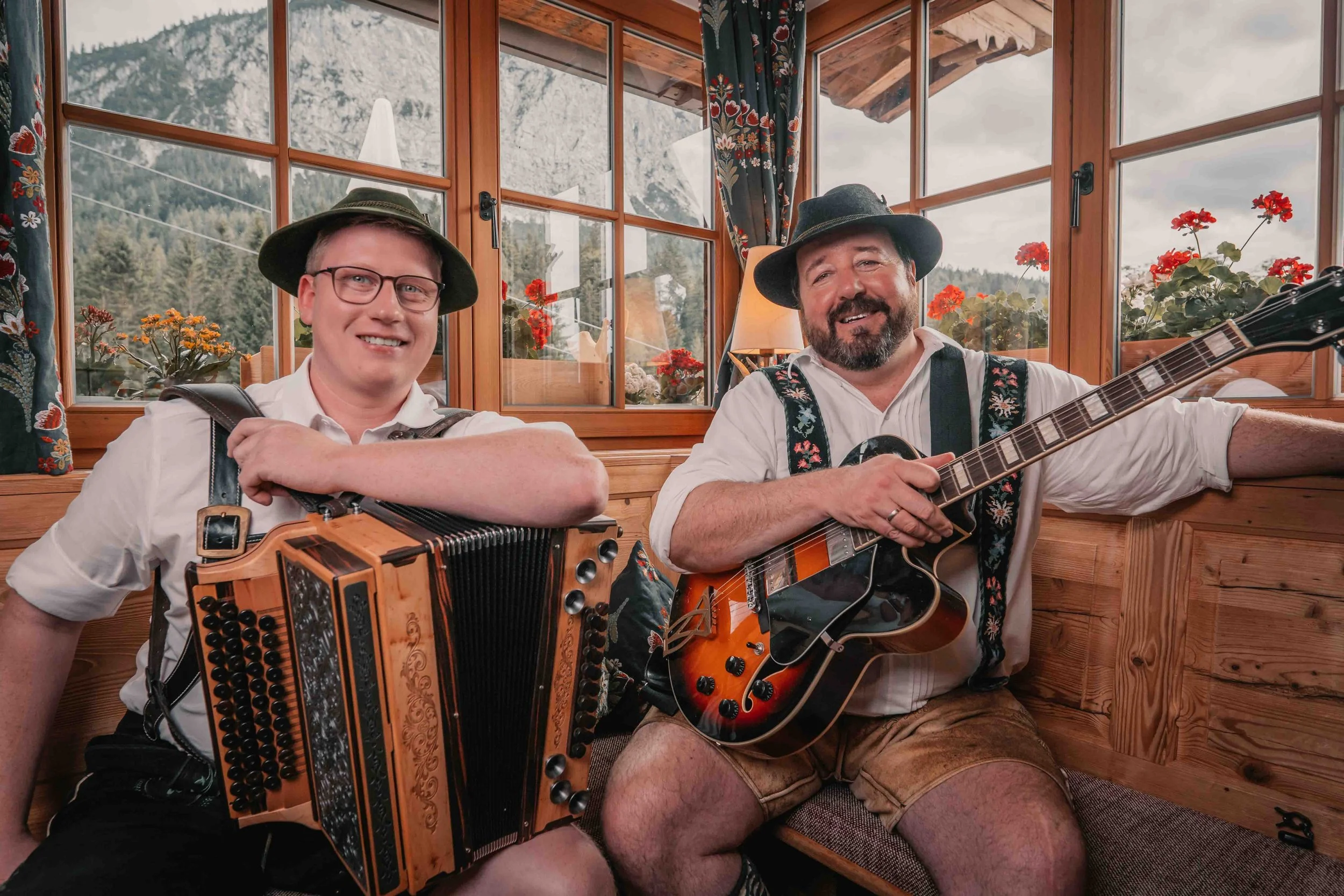 Zwei Männer in traditioneller Kleidung, die mit Musikinstrumenten in einem Holzhaus sitzen, mit Bergblick und Blumen im Hintergrund.