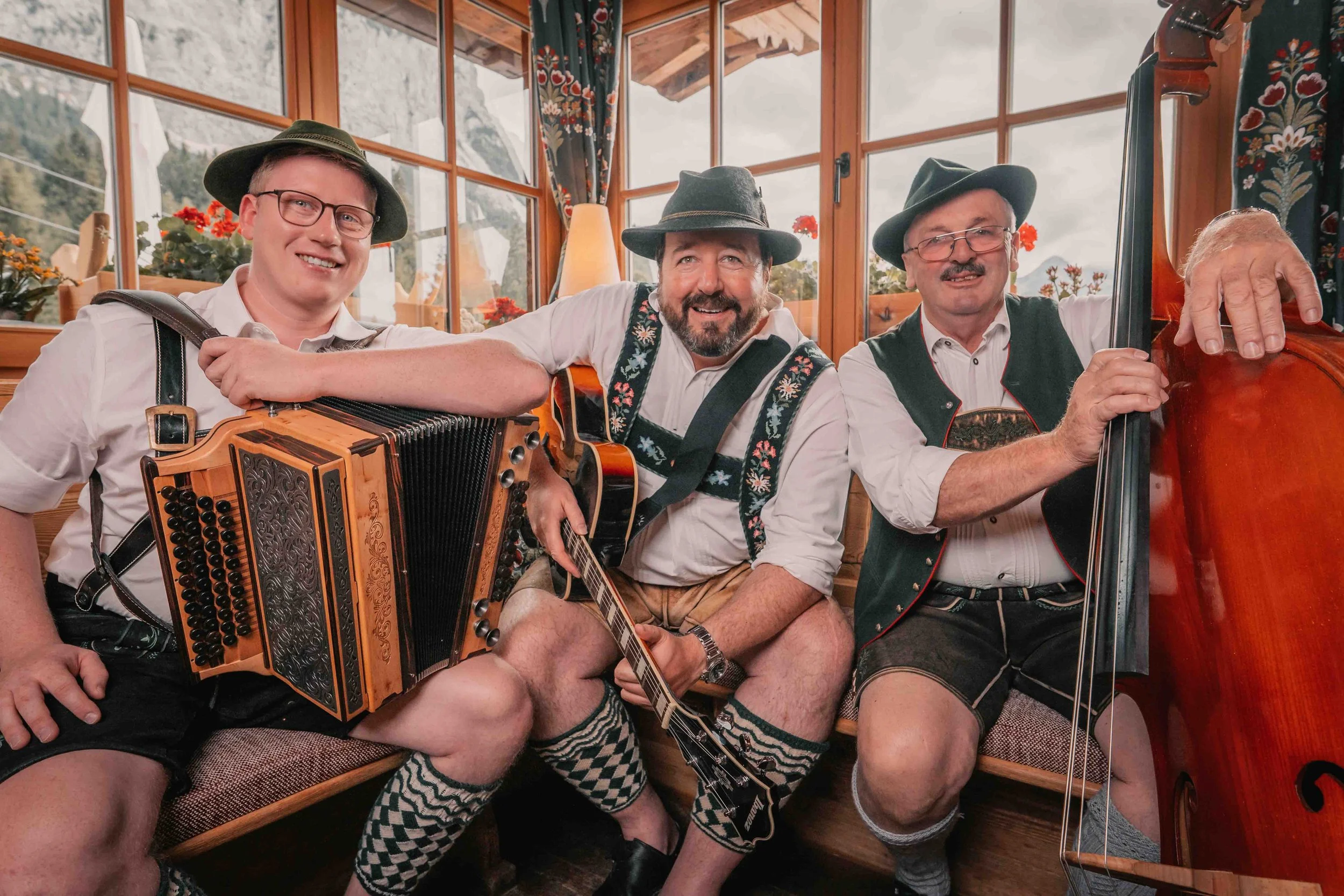 Drei Männer in traditioneller bayerischer Kleidung spielen Musik in einem Holzhaus mit großen Fenstern, umgeben von Bergen.