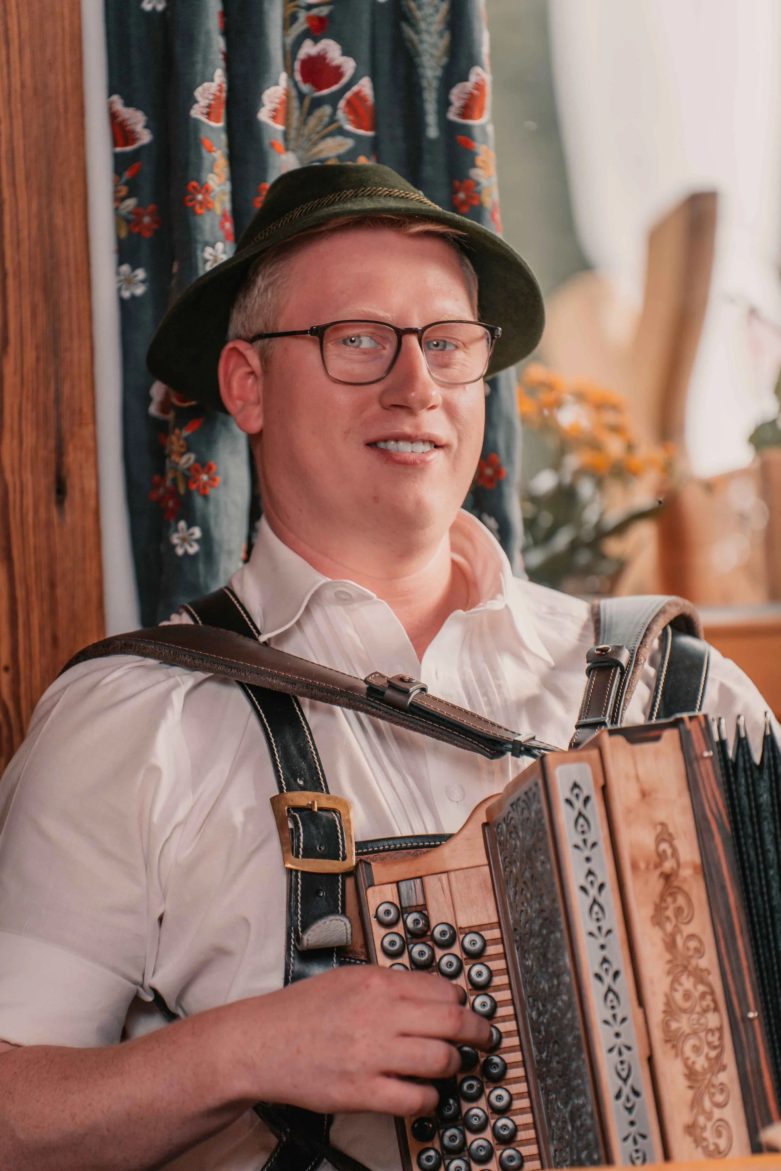 Ein Mann trägt eine Weißes Hemd, eine schwarze Lederhose, eine grüne Hut, spielt auf einem Zitter und lächelt.