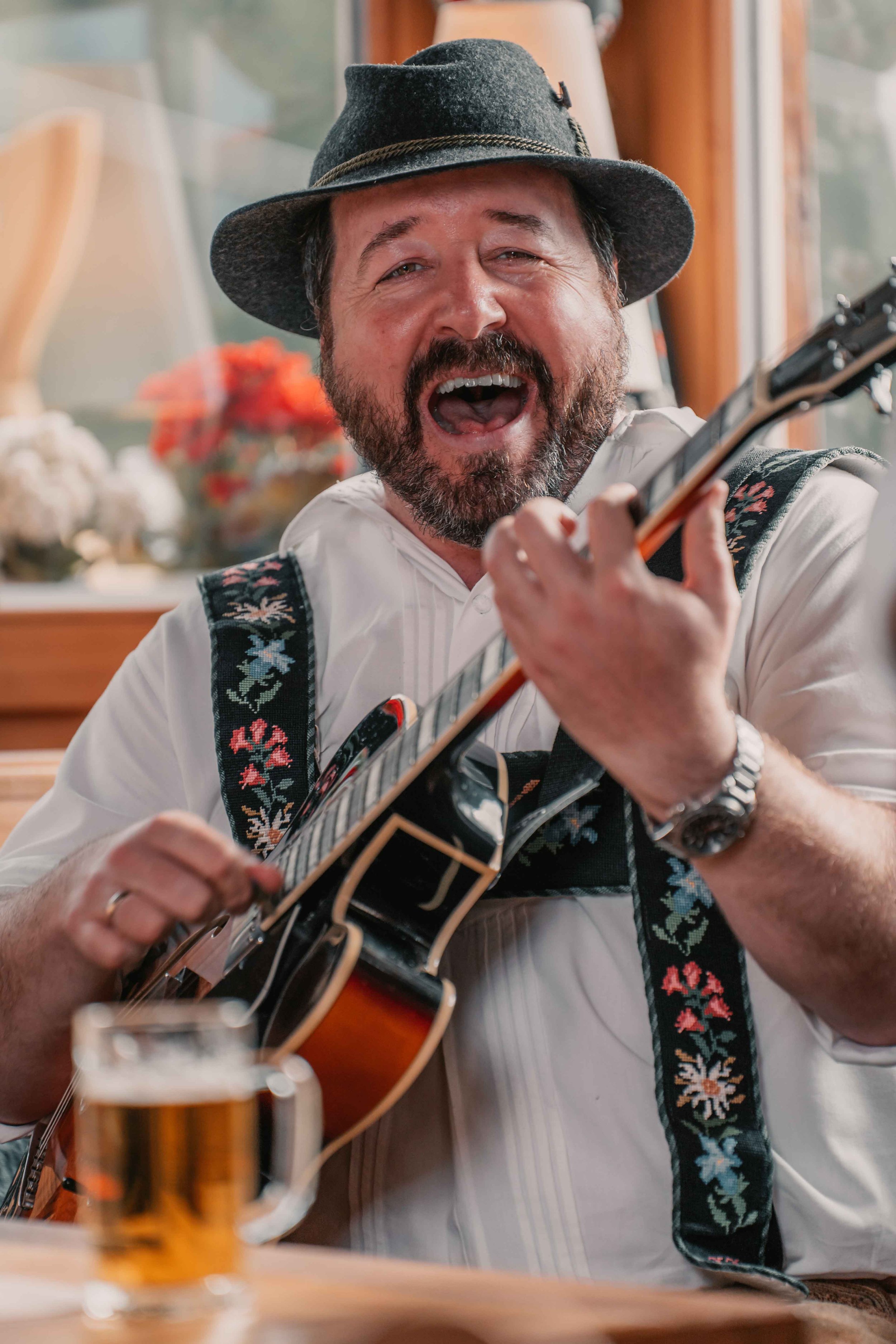 Ein lachender Mann trägt einen traditionellen Hut, spielt Gitarre und singt in einer gemütlichen Umgebung mit einem Bier auf dem Tisch.
