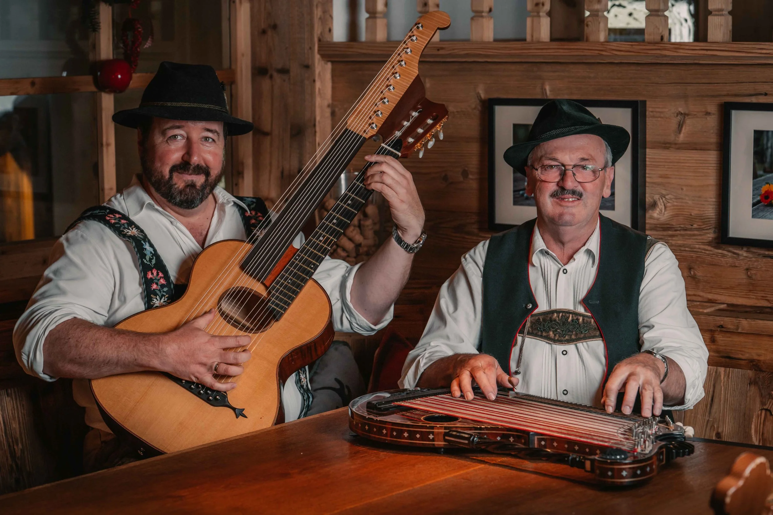 Zwei Männer in traditioneller deutscher Kleidung spielen Musik in einem Holzhaus, einer mit Gitarre, der andere mit Zither, beide tragen Trachtenhüte.