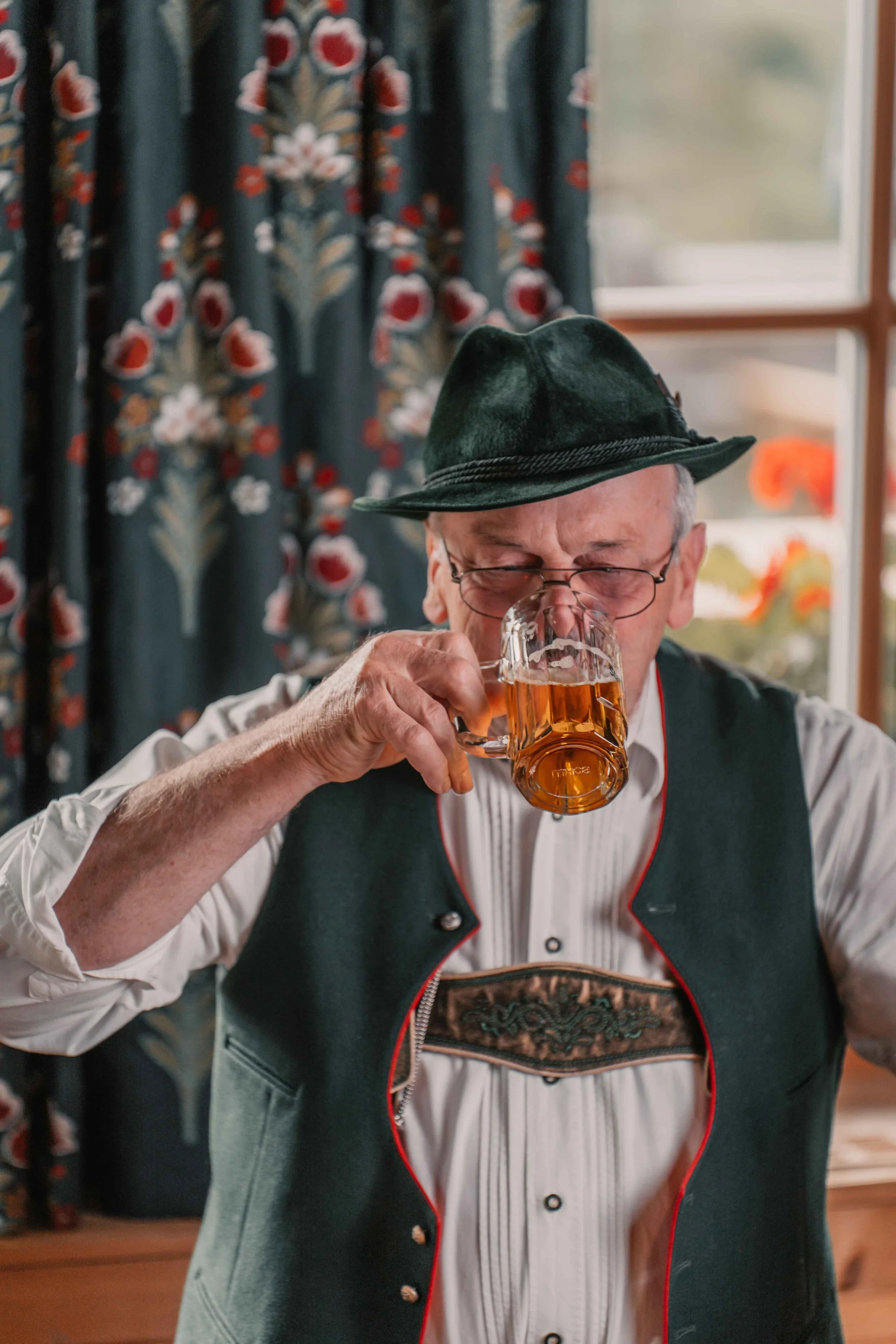 Ein älterer Mann in traditioneller bayrischer Tracht, der ein Bierglas hebt und trinkt, vor einem Fenster mit bunten Vorhängen.